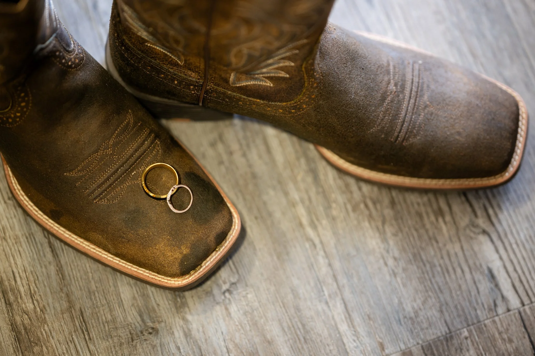 A pair of cowboy boots on a wooden floor with two wedding rings inside one of the boots.