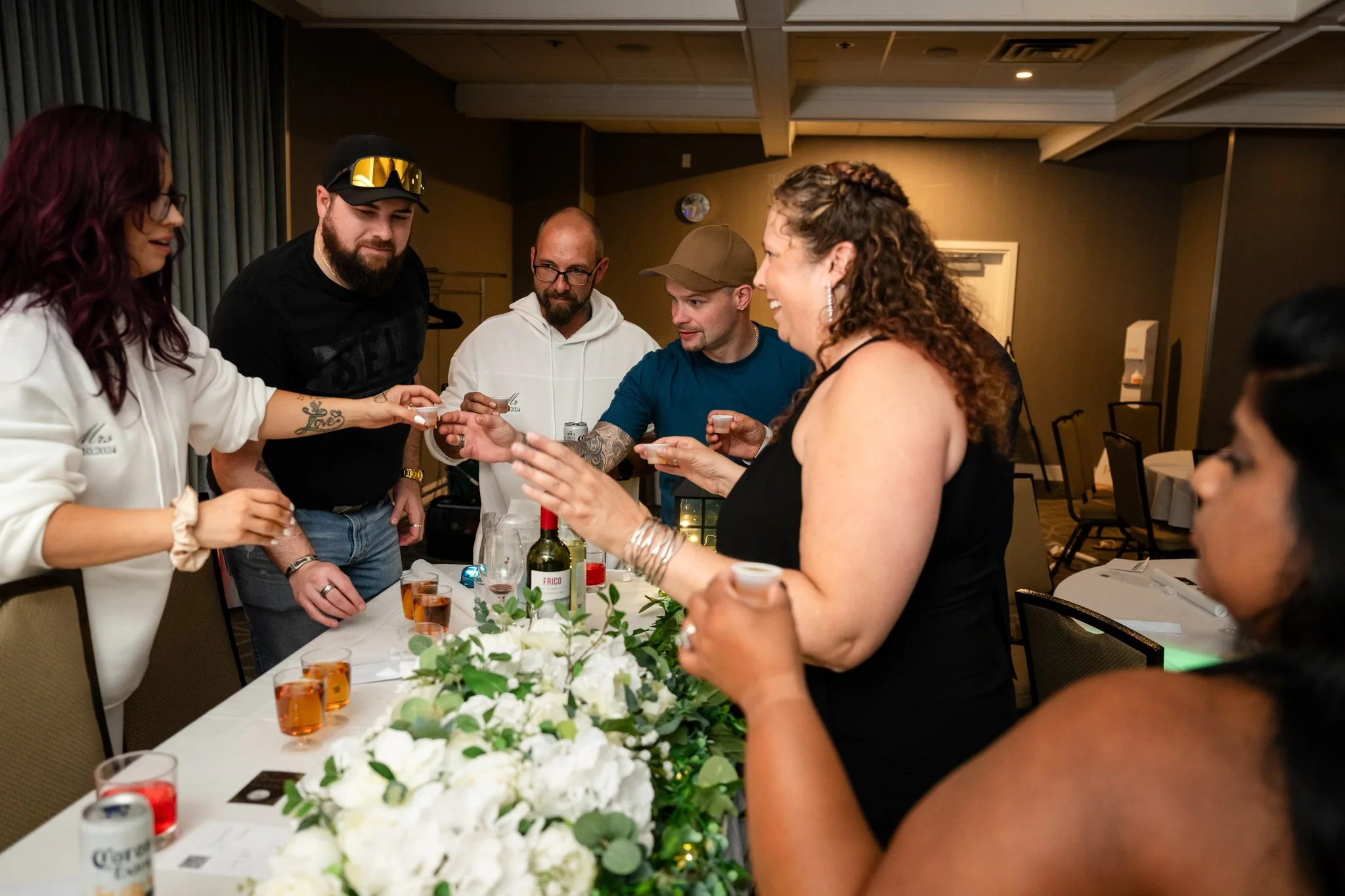 Group of six people gathered around a table, exchanging small cups, possibly during a celebration or toast. There are drinks, a wine bottle, and a floral centerpiece on the table.