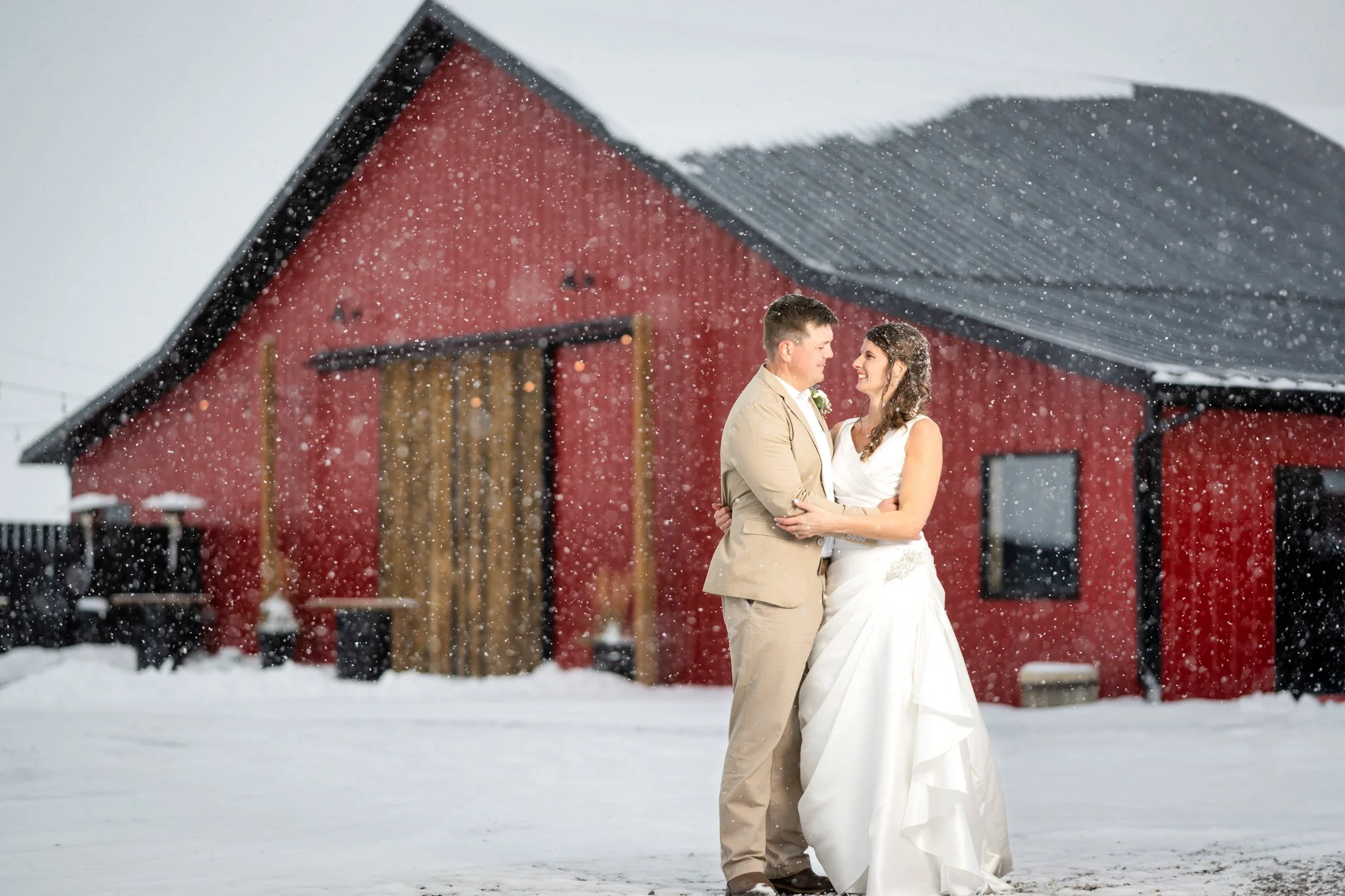 Bride and groom in wedding attire embracing outdoors in snowy weather, with a red barn in the background.