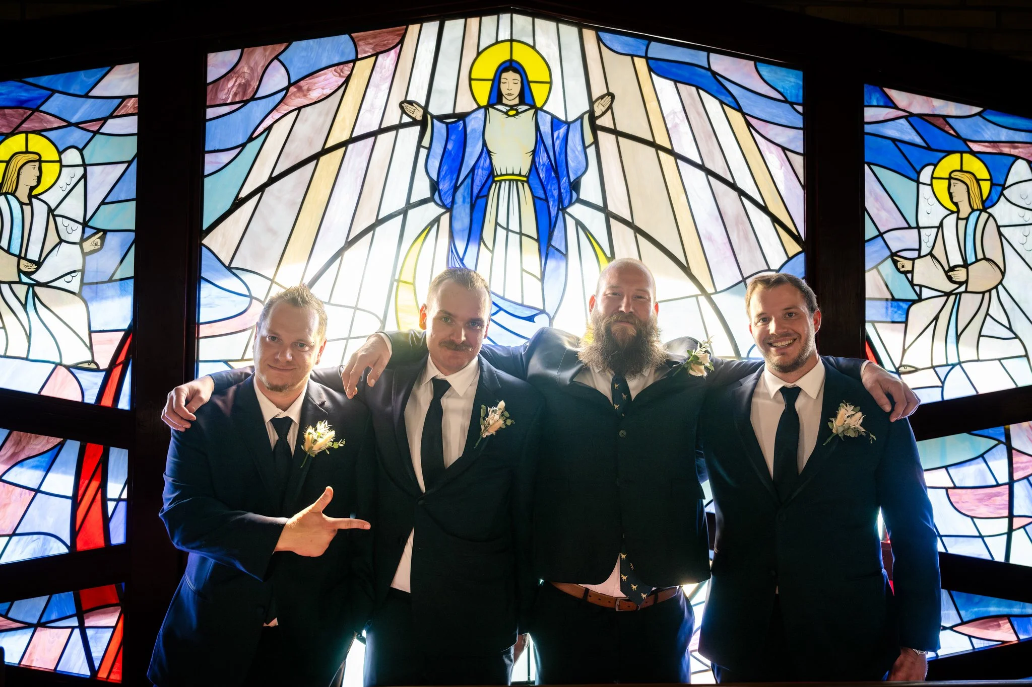 Four men dressed in suits with boutonnières, standing in front of a stained glass window depicting Jesus with open arms surrounded by angels.