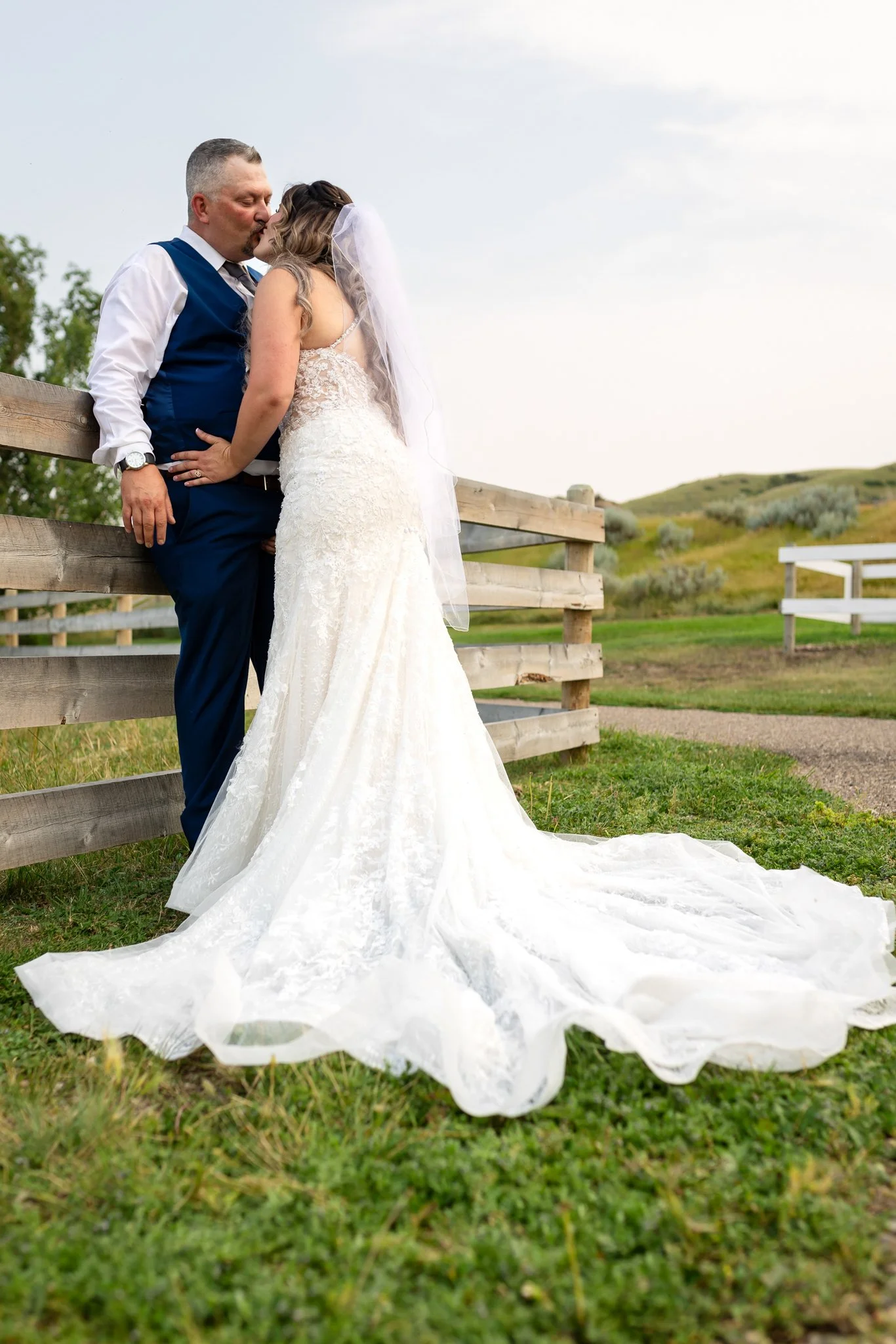 A bride and groom kissing outdoors on a grassy field, with a wooden fence and rolling hills in the background. The bride is in a white lace wedding dress with a long train and veil, while the groom is in a blue vest and white shirt.