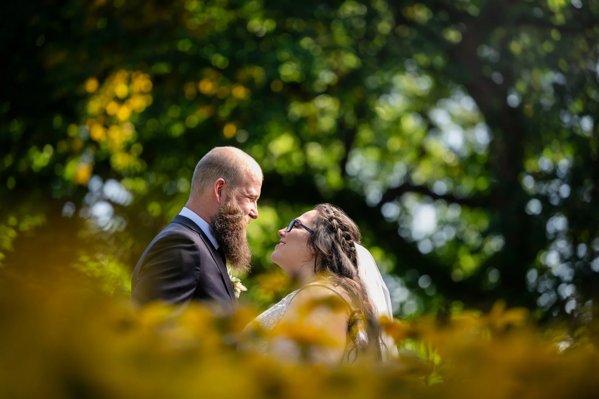A bride and groom standing closely together outdoors, looking into each other's eyes, with trees and greenery in the background.