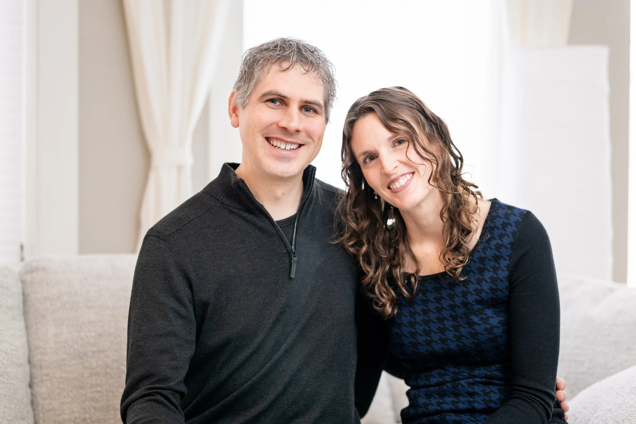 A smiling man and woman sitting on a light-colored sofa in a well-lit room, looking at the camera.