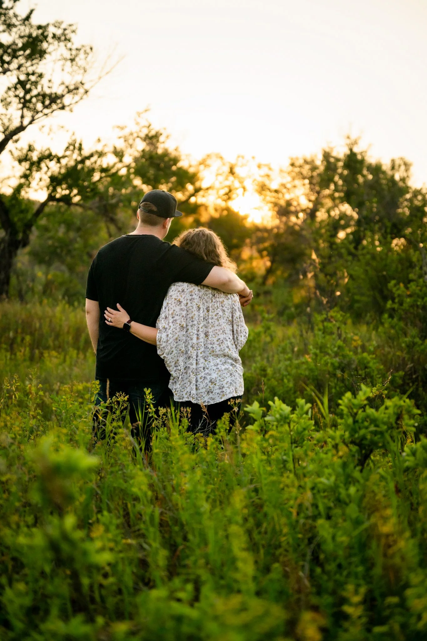 A couple walking arm in arm through a green field at sunset, with trees in the background.