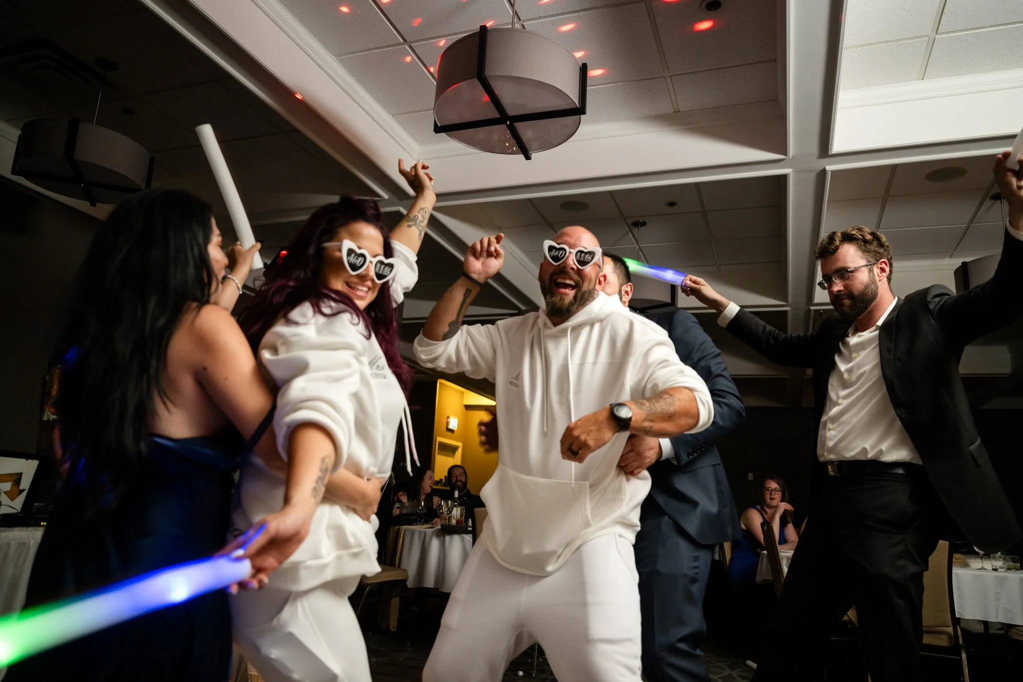 People dancing and celebrating at a party or wedding reception, wearing sunglasses, holding light sticks, and smiling.