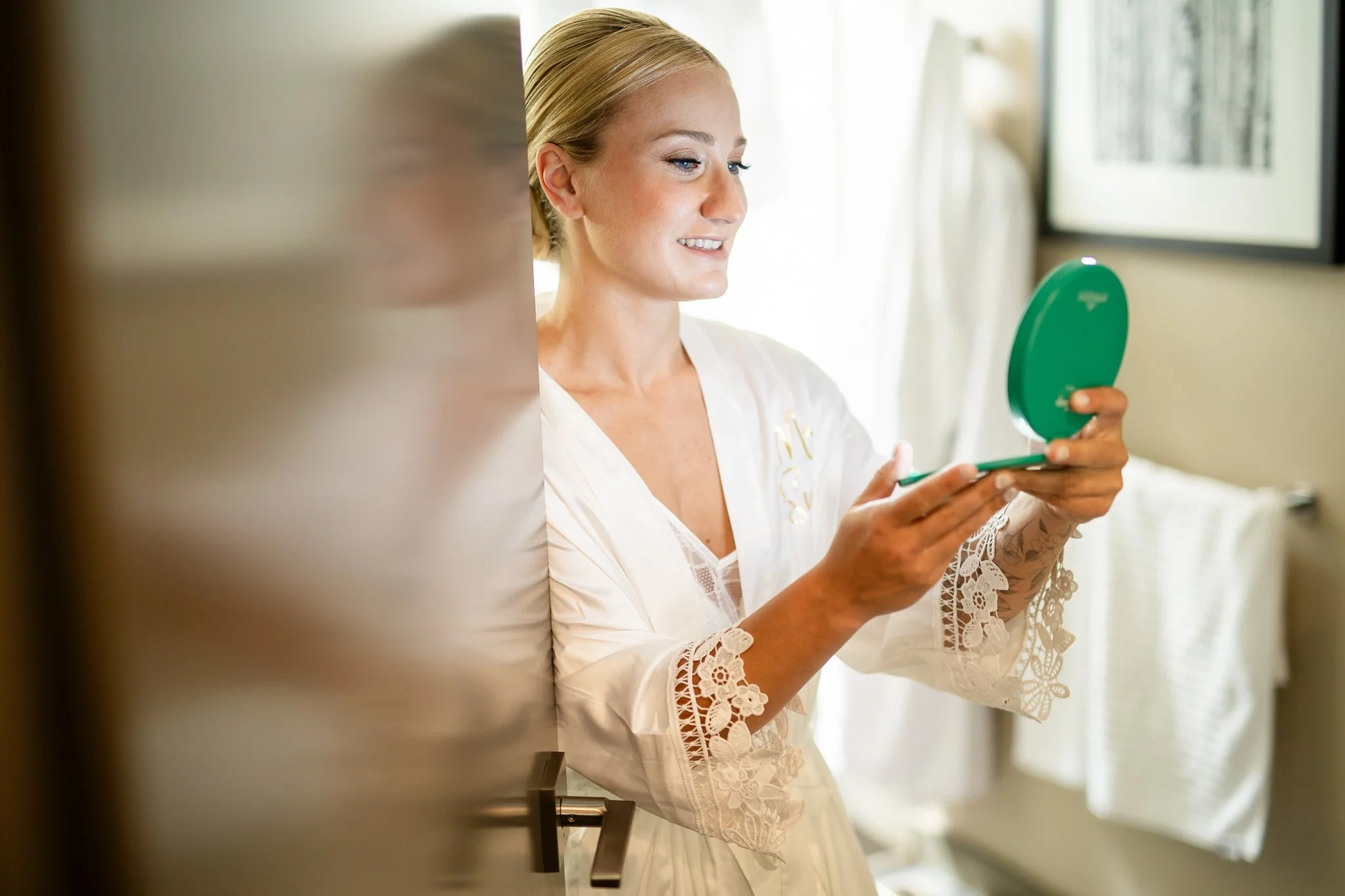 A woman with blonde hair looking in a mirror while holding a compact mirror, smiling, in a bright room with natural light.