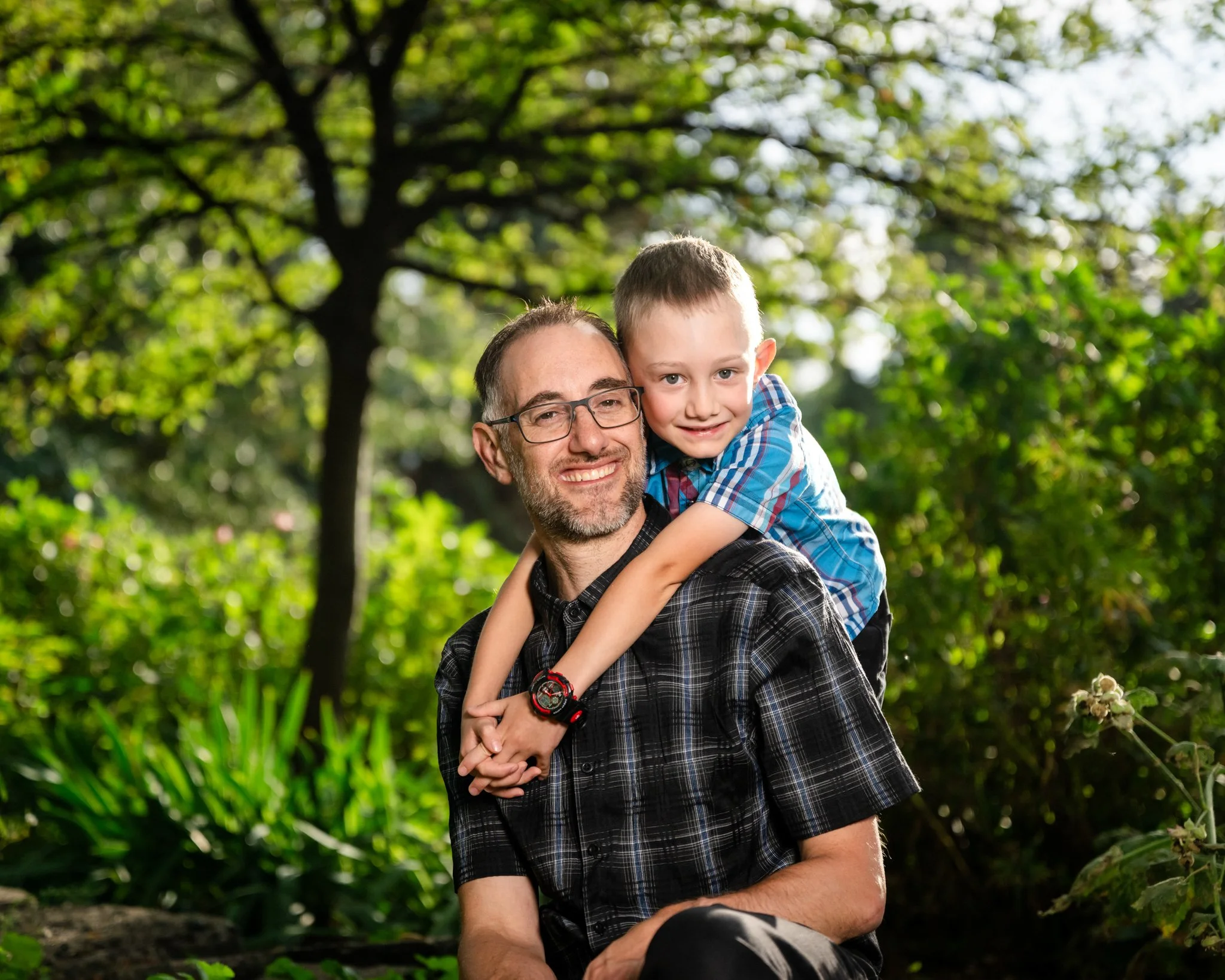 A smiling man with glasses sitting outdoors with a young boy on his shoulders, surrounded by greenery.