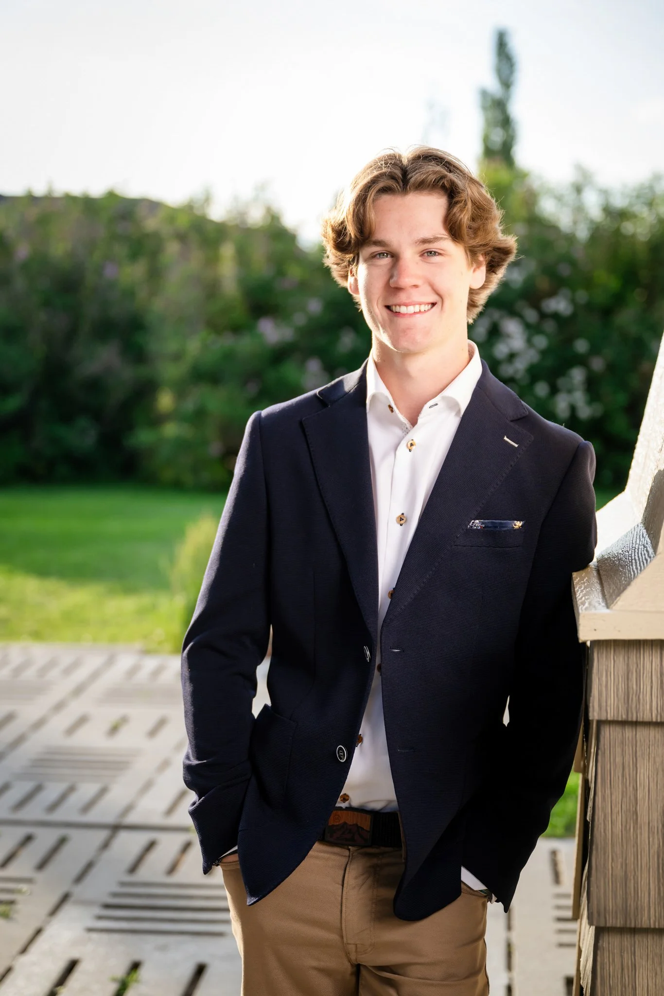 A young man in a navy blazer and white shirt standing outdoors on a wooden deck, smiling at the camera with a background of green trees and grass.