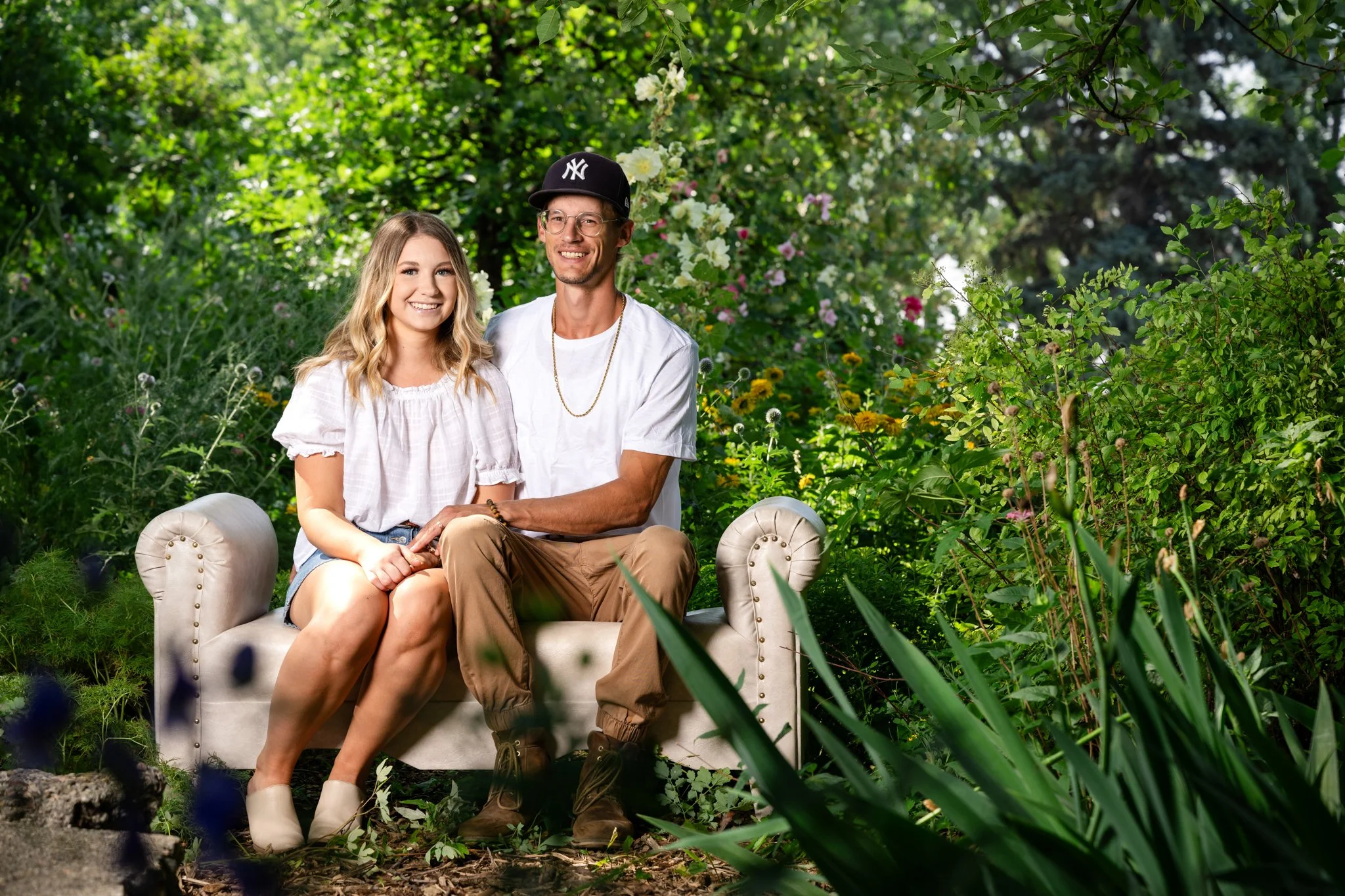 A young woman and a man sitting on a light-colored tufted sofa outdoors in a lush garden with green foliage and colorful flowers, smiling for the camera.