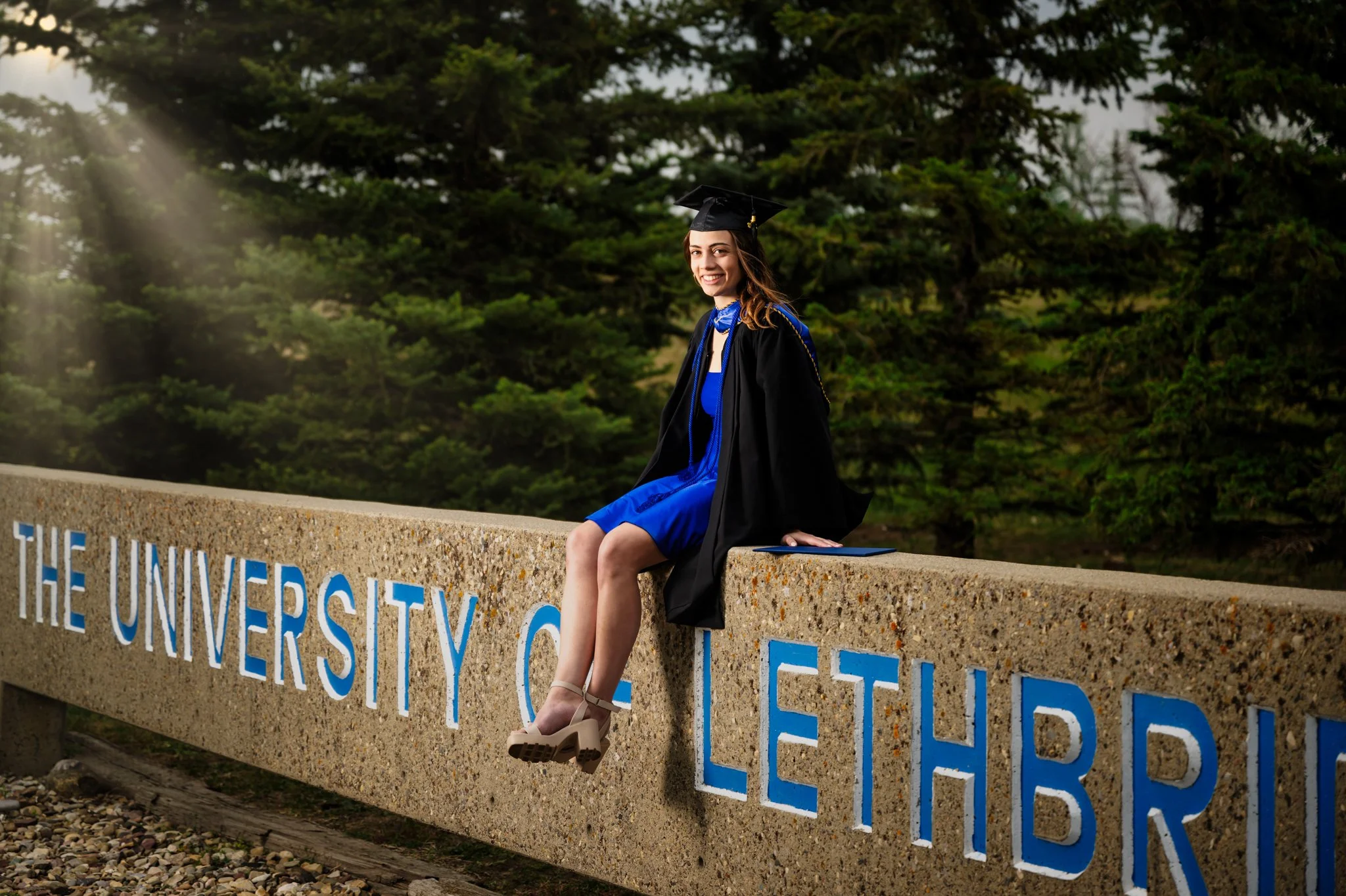 A smiling young woman in a blue dress and graduation gown with cap, sitting on a concrete ledge at the University of Lethbridge, outdoors with trees in the background.