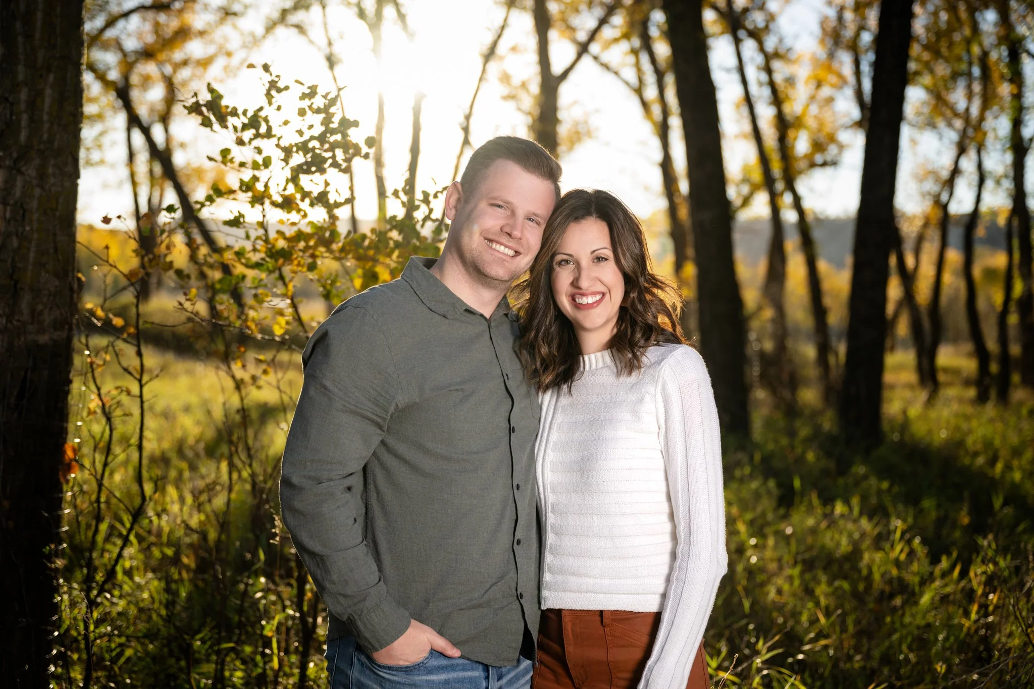 A smiling man and woman standing together outdoors in a wooded area during autumn, with sunlight streaming through the trees behind them.