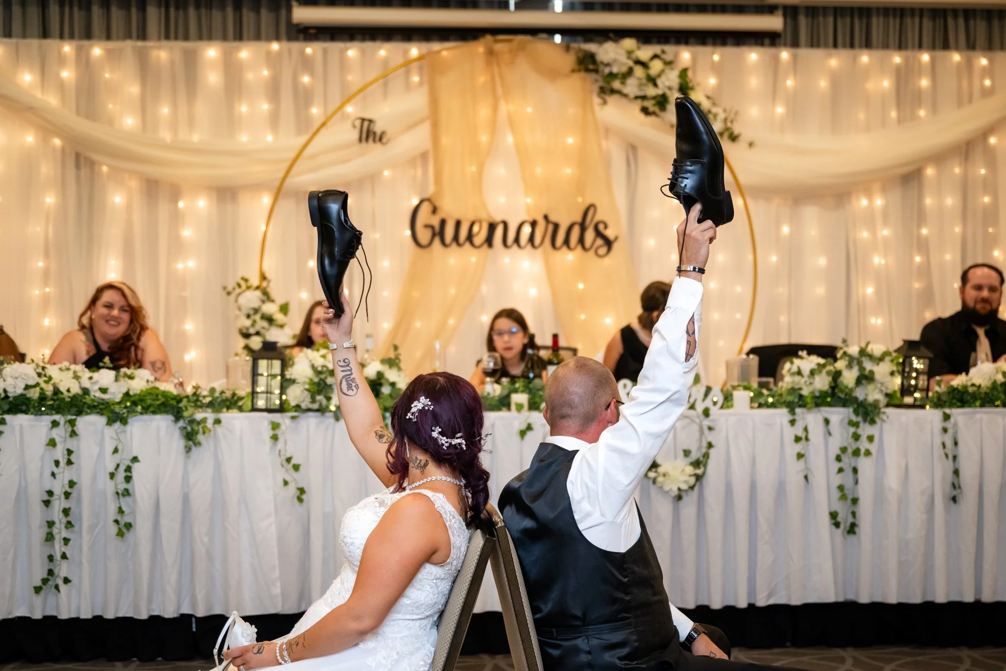 A newlywed couple sitting back-to-back on chairs named 'Groom' and 'Bride' with their arms raised, holding black dress shoes in the air during a wedding reception. The bride has purple hair and tattoos, and the groom is wearing glasses and a vest. Th
