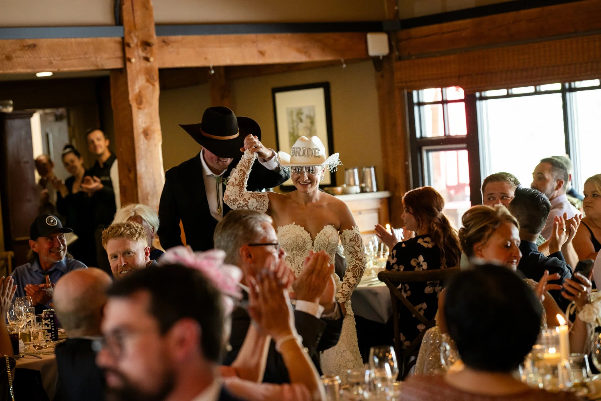 Bride in a white lace wedding dress wearing a 'bride' hat, smiling, as guests sit at tables clapping and celebrating in a rustic wooden venue.