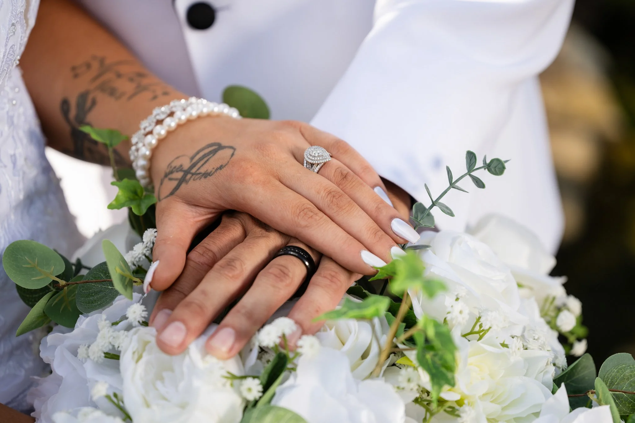 Close-up of a bride's hand with wedding rings, resting on a groom's hand, surrounded by white flowers and greenery.