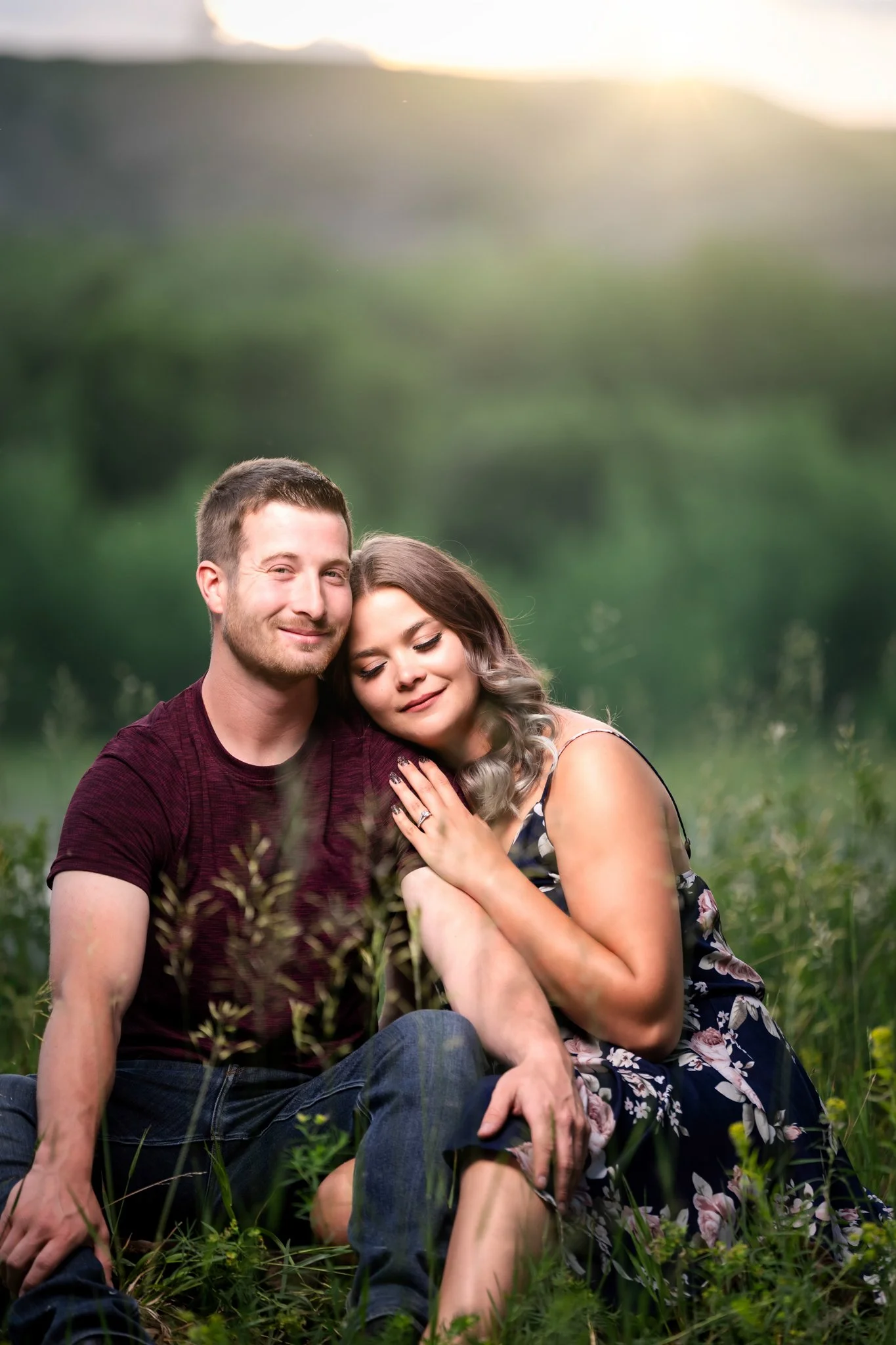 A couple sitting in a grassy field embracing each other, with a blurred green landscape and sunlight in the background.