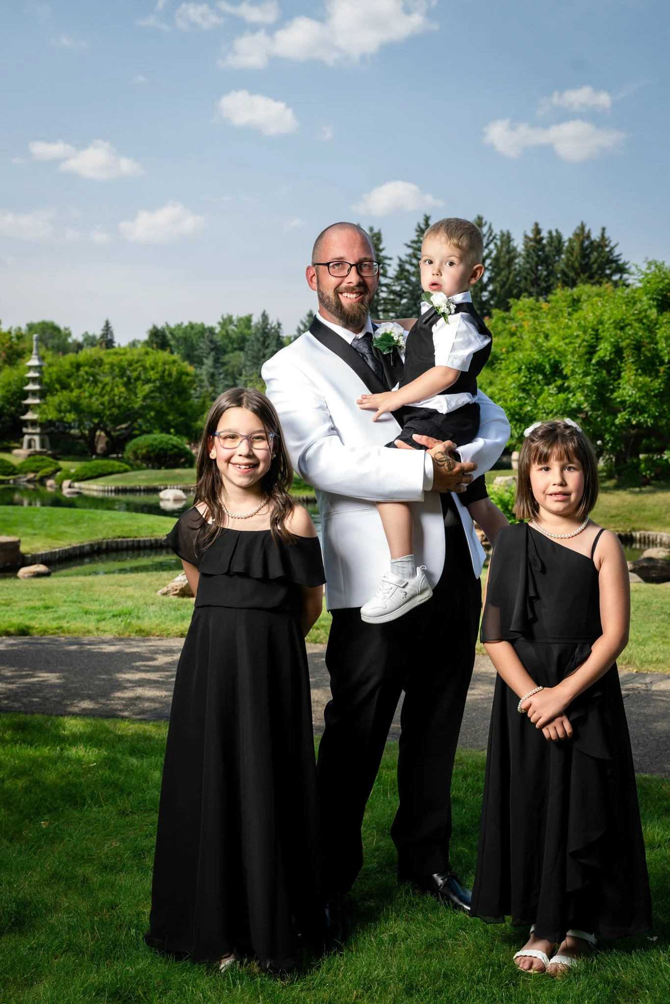 A man in a white tuxedo with black accents and glasses is holding a young boy in a black and white outfit. Two young girls in black dresses and pearl necklaces stand beside them in a garden with greenery, a pond, and a pagoda in the background.