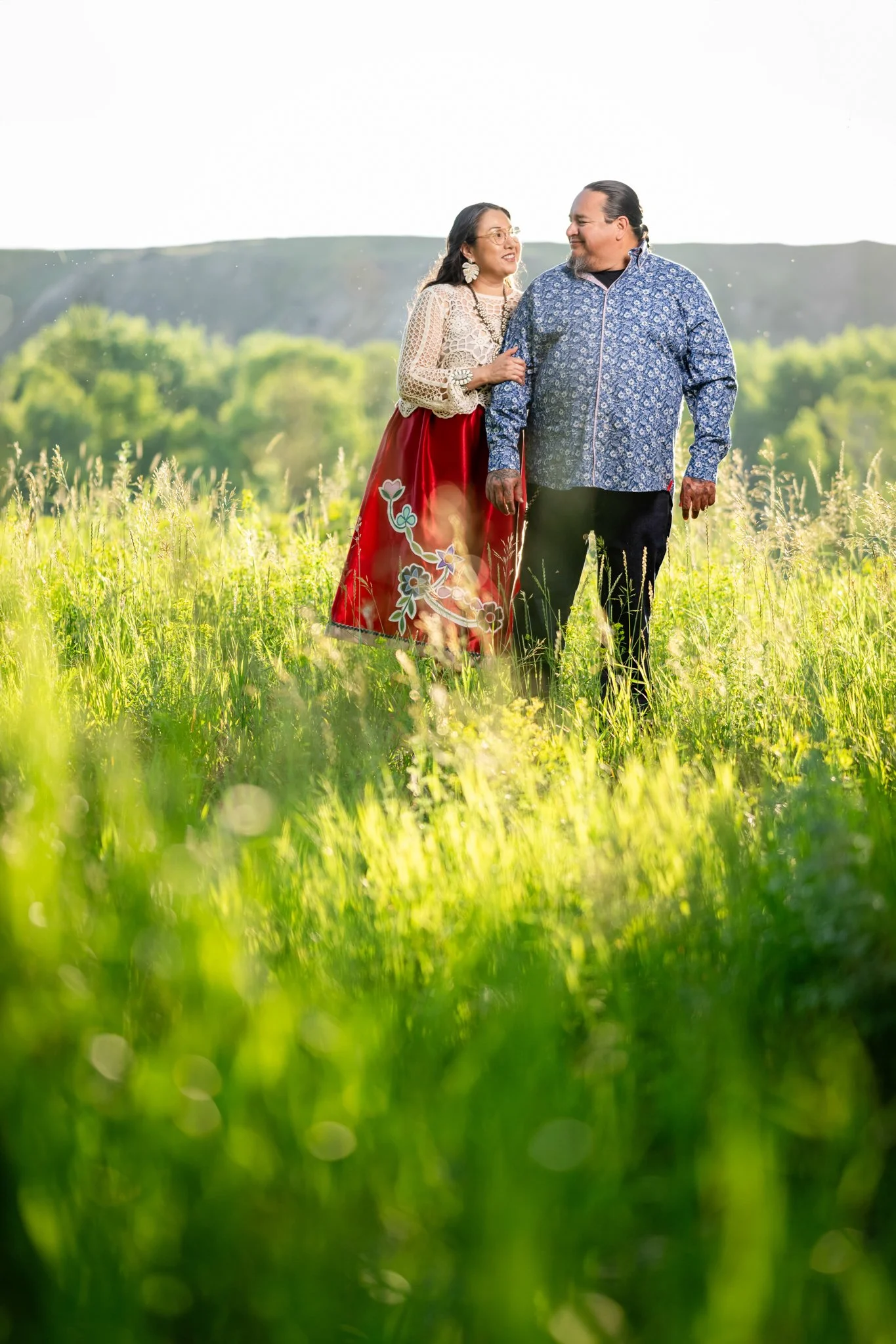 A man and woman walking in a lush green field during daytime, smiling and holding each other, with trees and hills in the background.