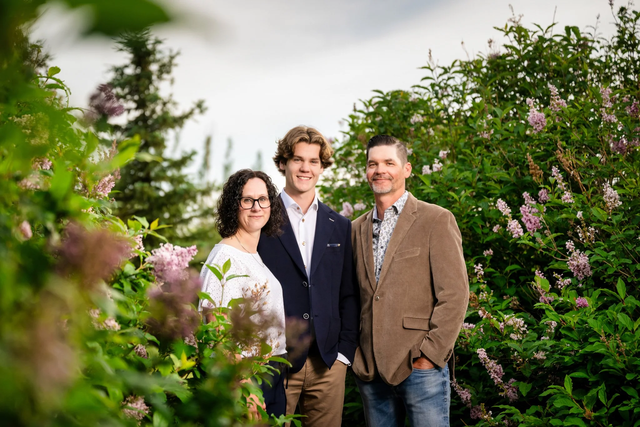 A family of three standing outdoors among flowering bushes. The woman has curly black hair and glasses, wearing a white blouse. The young man has wavy brown hair, wearing a navy blazer, white shirt, and tan pants. The man has short dark hair, a beard