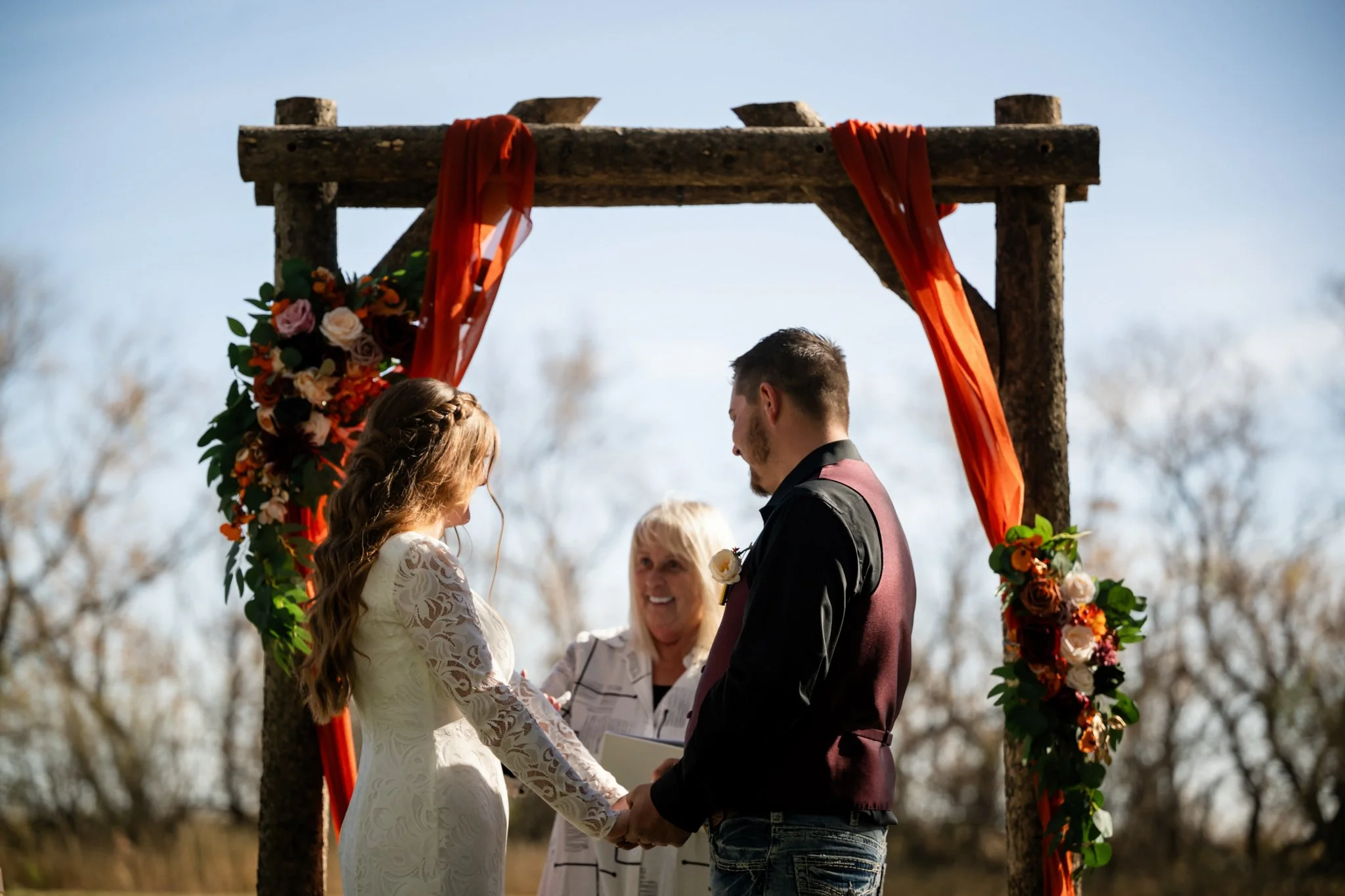 Couple getting married outdoors under a wooden arch decorated with red fabric and flowers, with an officiant officiating the ceremony.