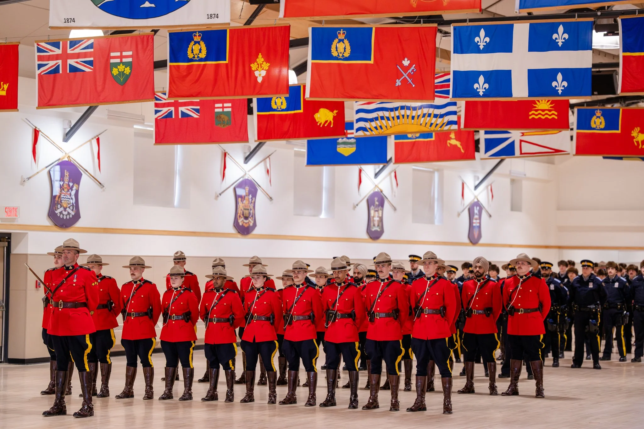 Ceremonial guards in red uniforms and khaki hats marching in formation inside a hall decorated with numerous flags and banners.