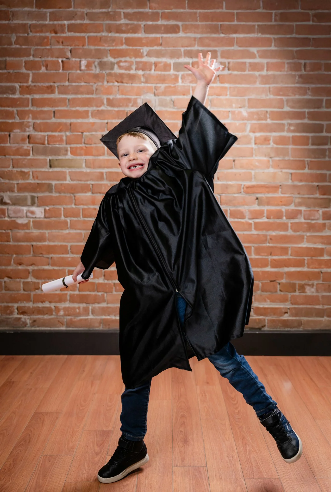A young boy in a graduation cap and gown, holding a diploma, joyfully dancing or celebrating with one arm raised, standing in front of a brick wall.