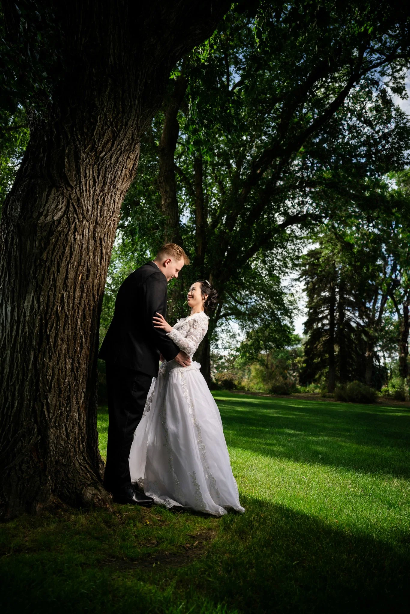 A bride and groom standing close together under a large tree in a lush, green park. The bride is wearing a white wedding gown, and the groom is in a black suit. They are smiling at each other.