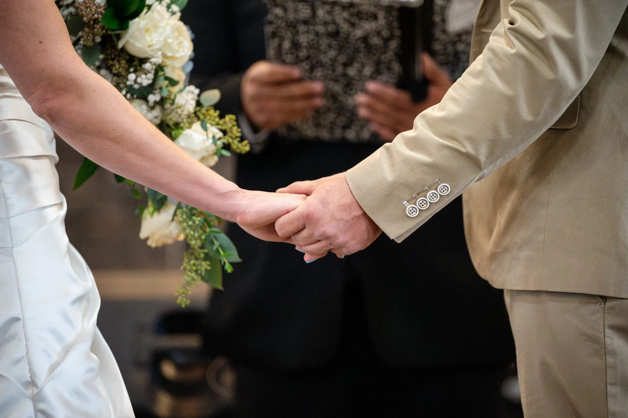 A couple holding hands during a wedding ceremony, with a bouquet of white flowers visible to the left.