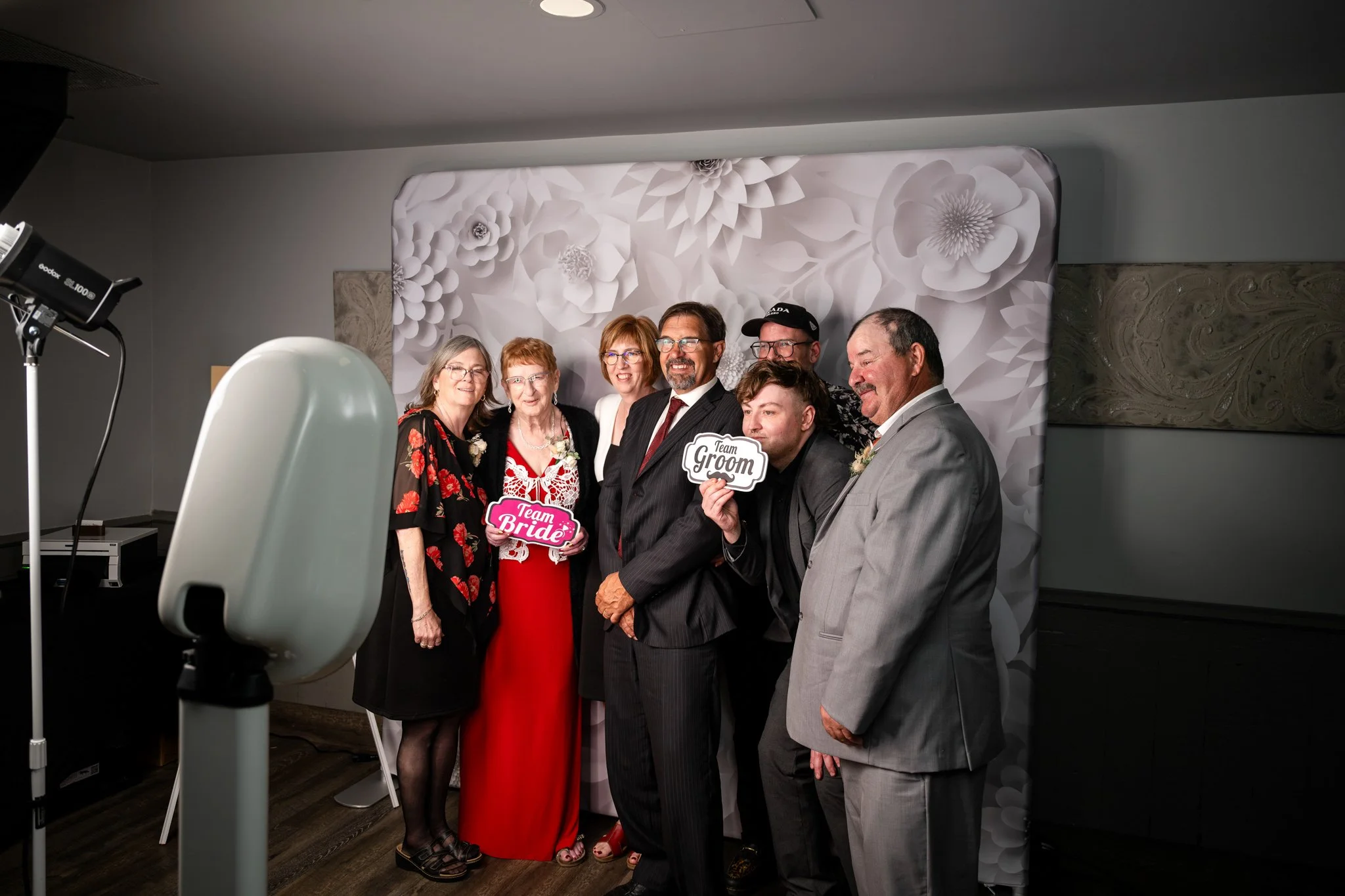Group of people at a wedding photo booth, holding signs that say 'Team Bride' and 'Team Groom', with a white floral backdrop.