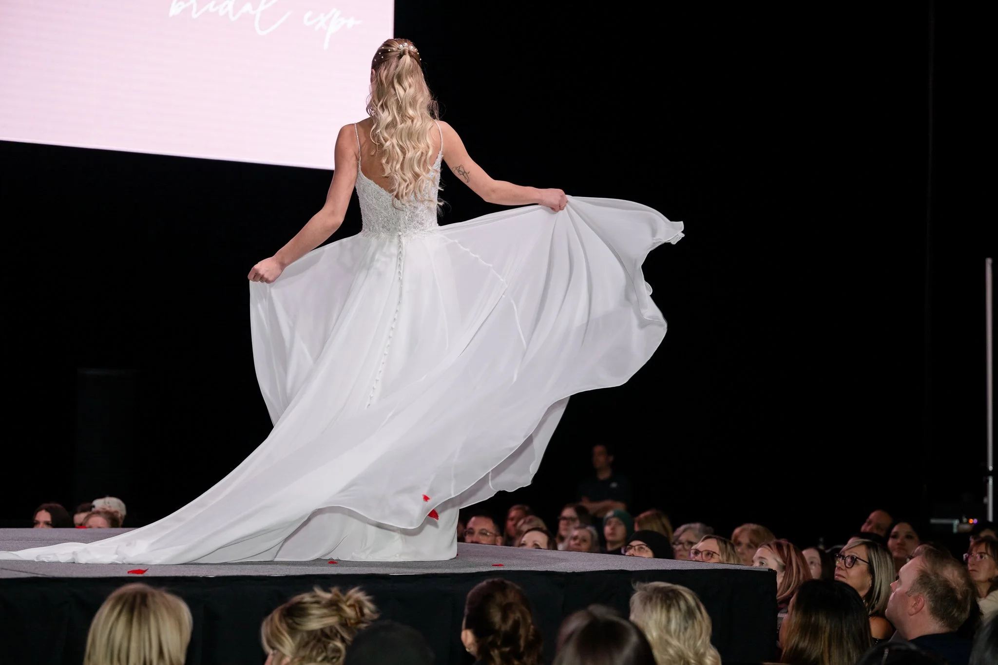 A woman in a white wedding dress walking on a runway at a fashion show with an audience watching.