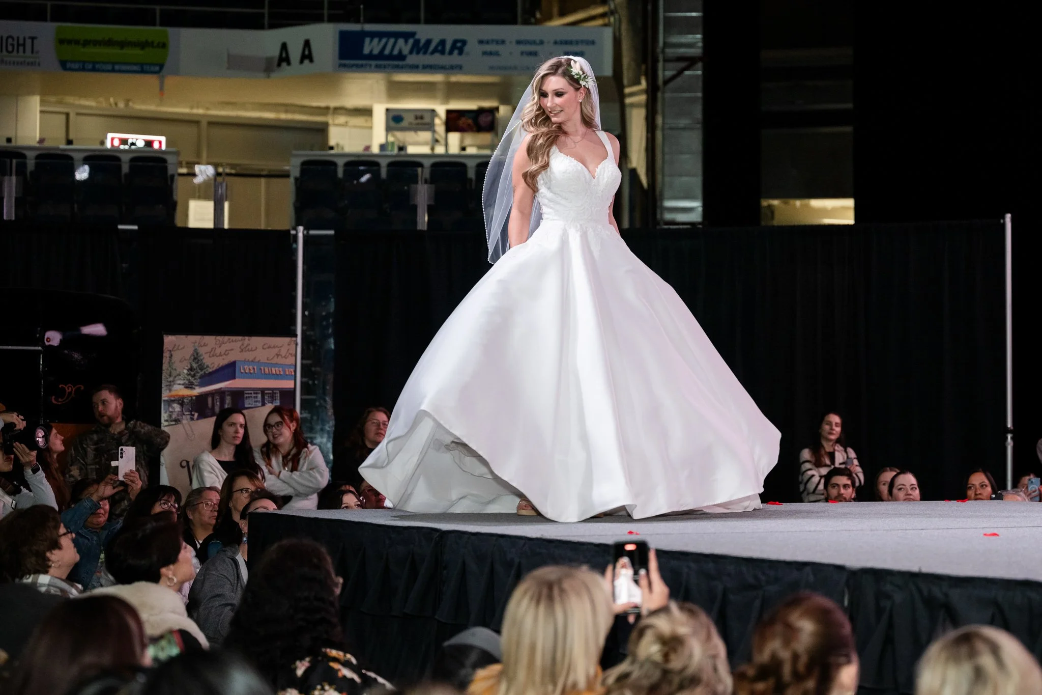 A woman in a white wedding dress and veil walking on a runway with an audience watching.