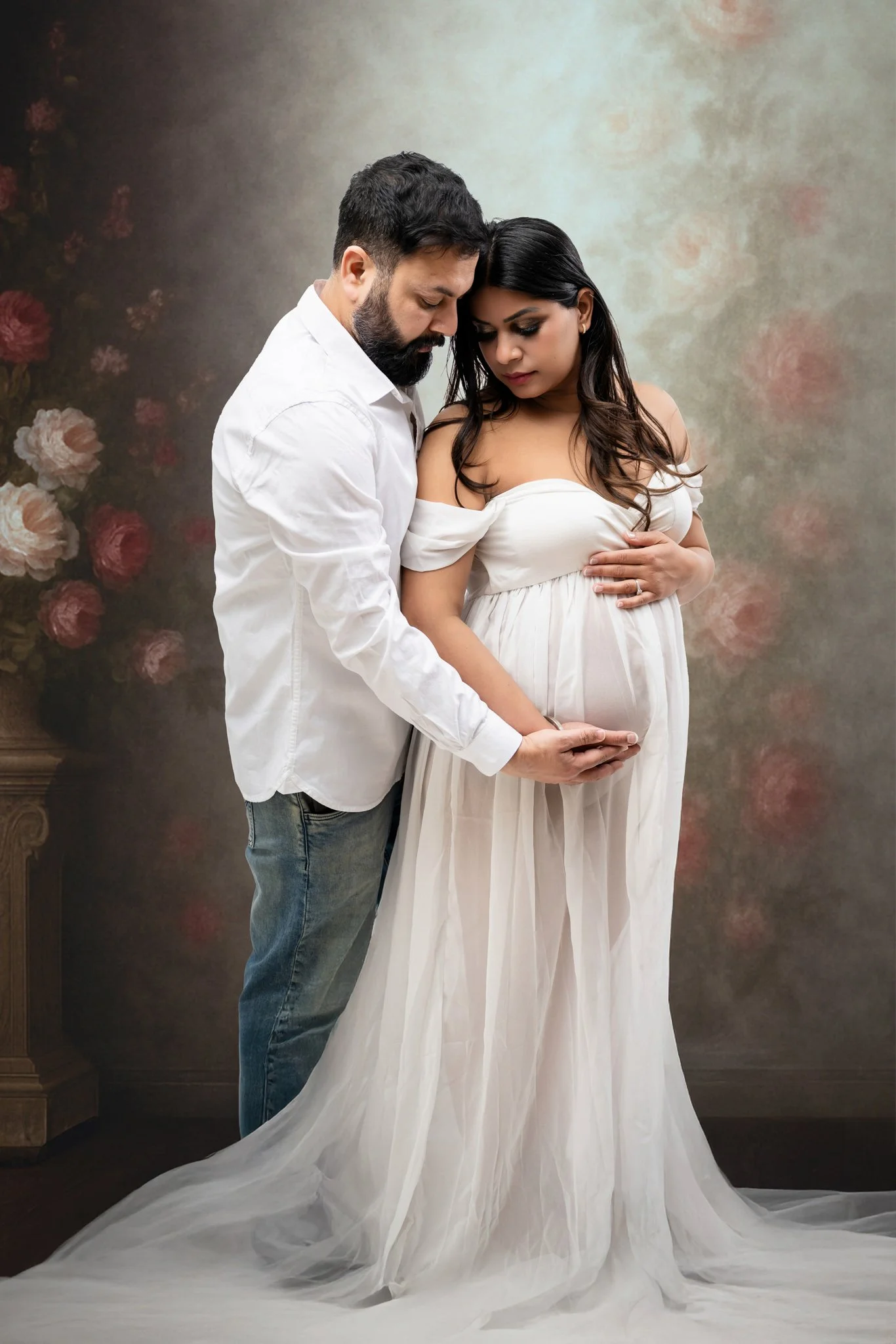 A pregnant woman in a white dress with her hand over her belly, standing next to a man in a white shirt who is gently holding her belly with both hands, with a floral backdrop.
