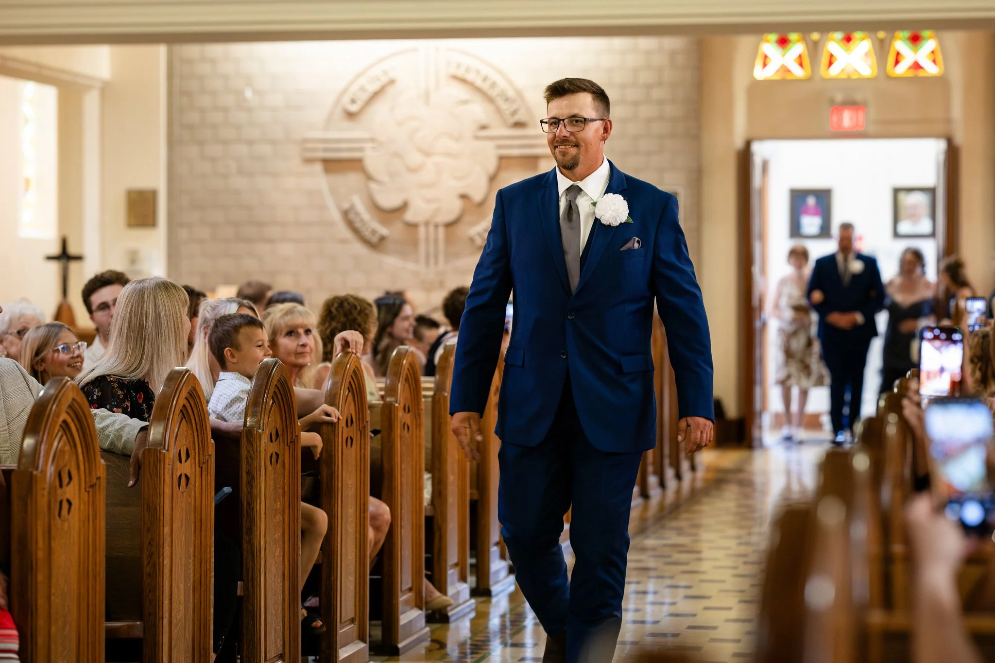 A groom in a blue suit walking down the aisle at a wedding ceremony in a church, with guests seated on either side watching and taking photos.