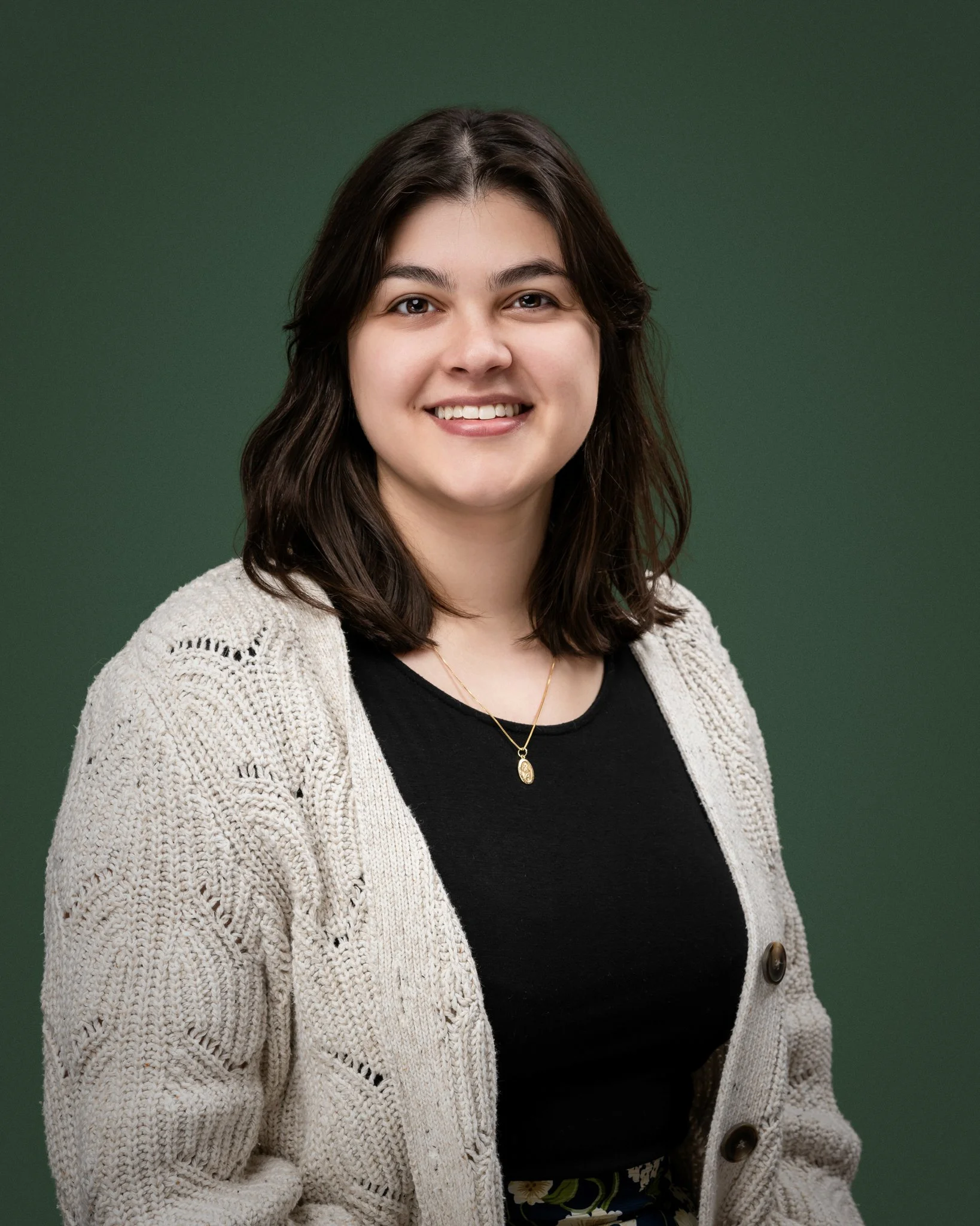 A young woman with shoulder-length dark hair, wearing a beige knitted cardigan over a black top, smiling at the camera against a green background.