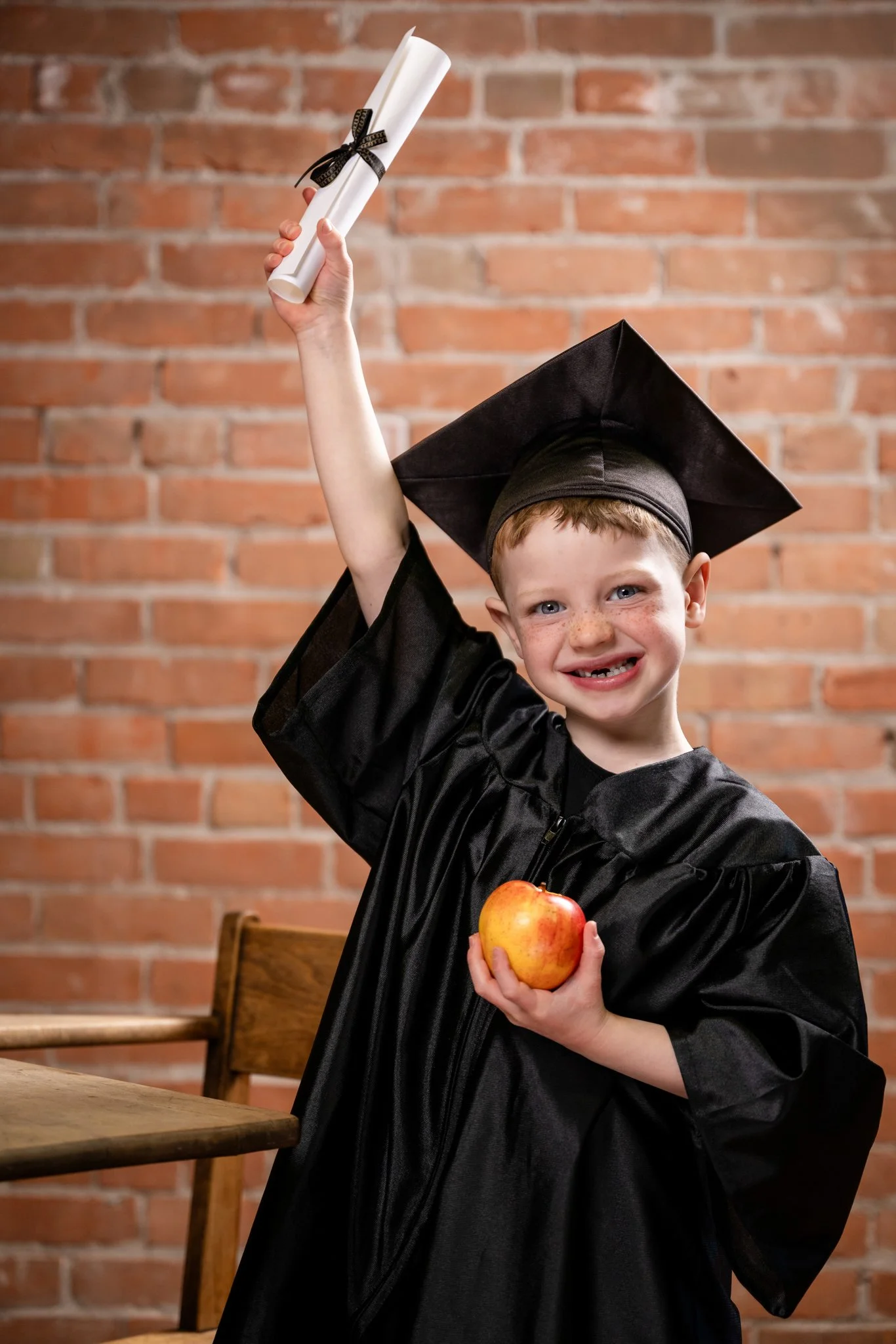 A young boy in a black graduation gown and mortarboard cap is smiling at the camera, holding an apple in his left hand and a rolled diploma tied with a black ribbon in his right hand against a brick wall background.