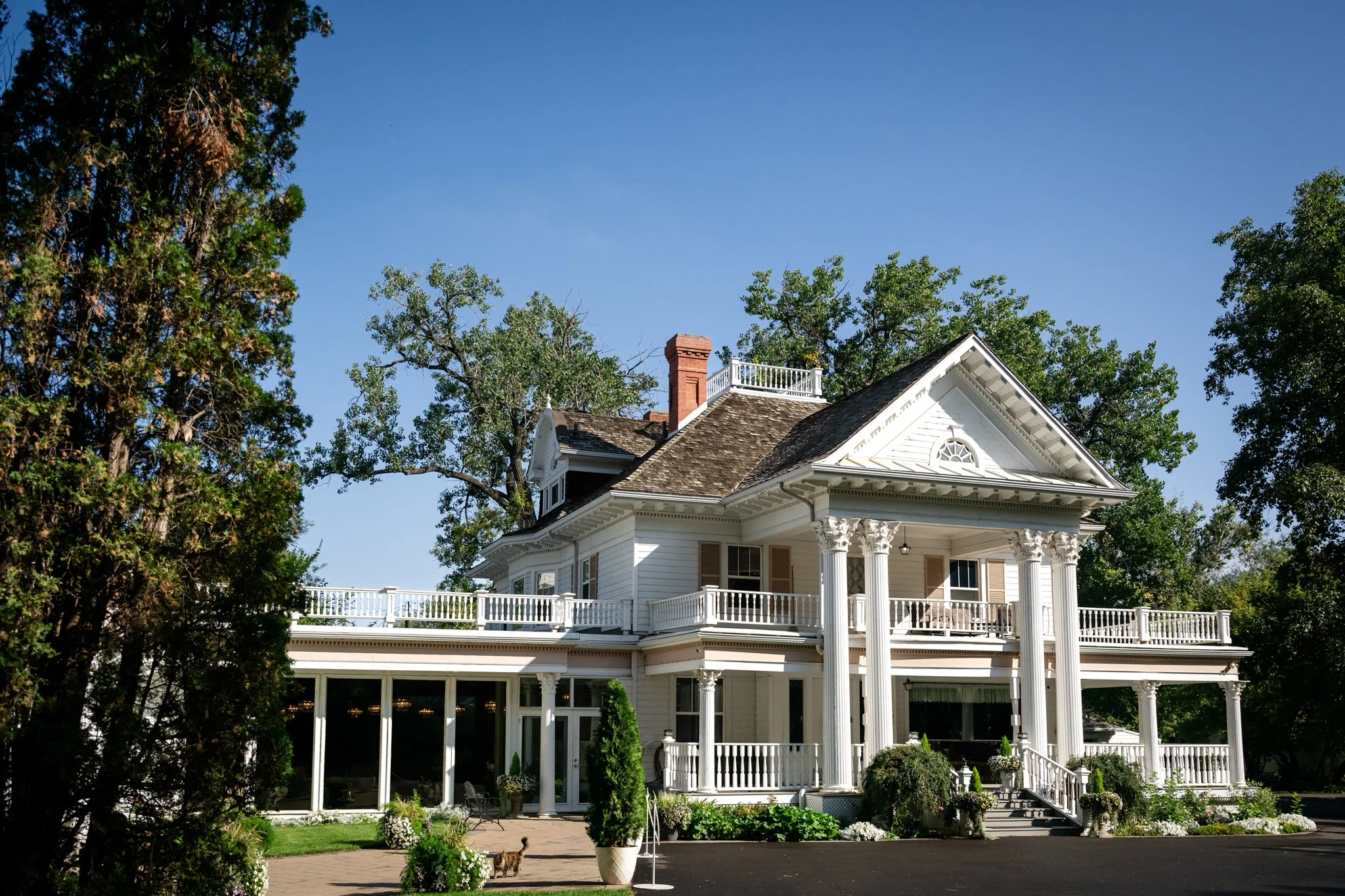 Large white historic house with columns, surrounded by trees and flowers, under a clear blue sky.