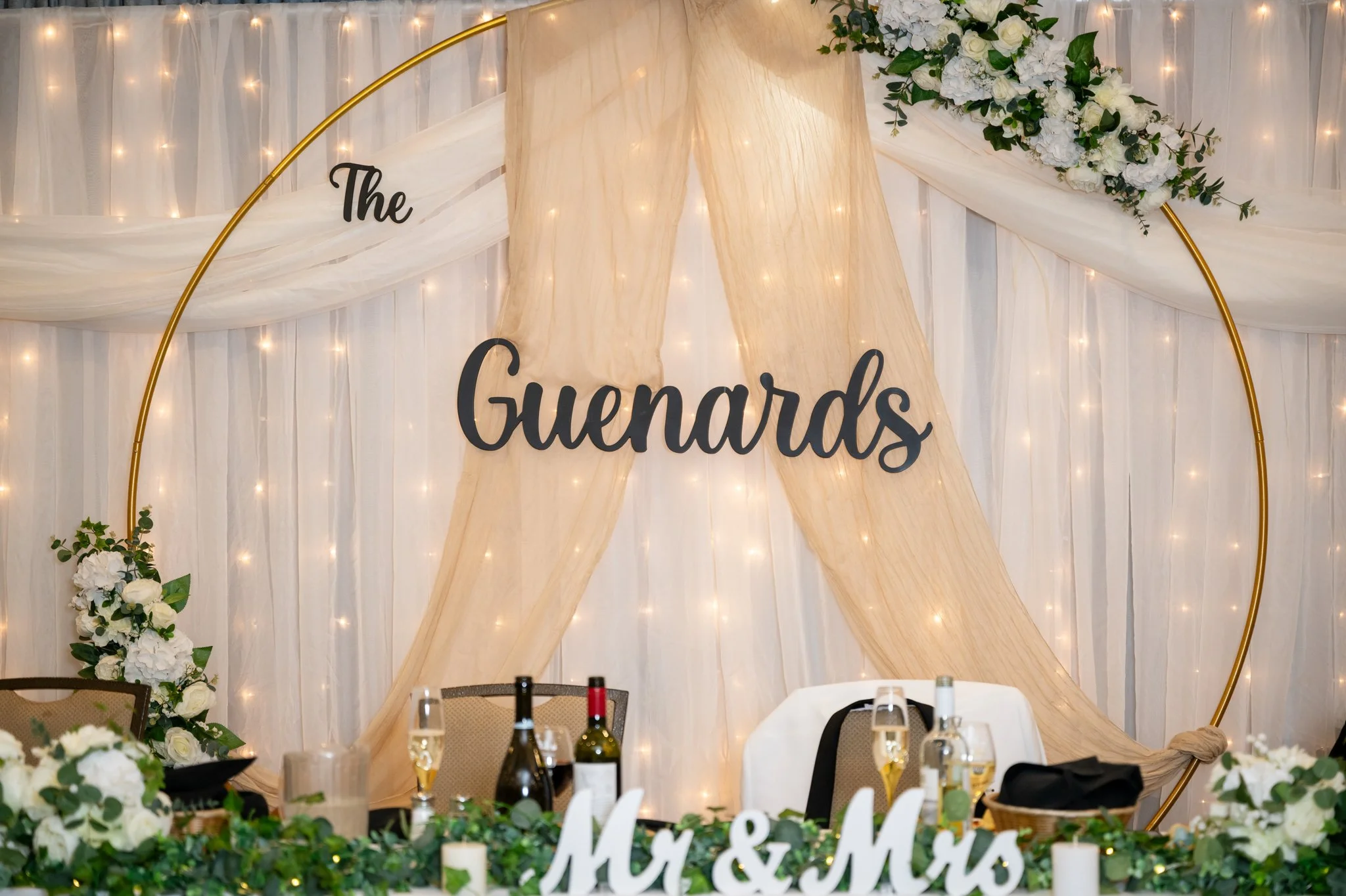 Wedding or celebration decor featuring a backdrop with white and cream drapes, fairy lights, floral arrangements, a large circle frame, and a sign that reads 'The Guenards'. There are tables with wine bottles, glasses, and a 'Mr & Mrs' sign.