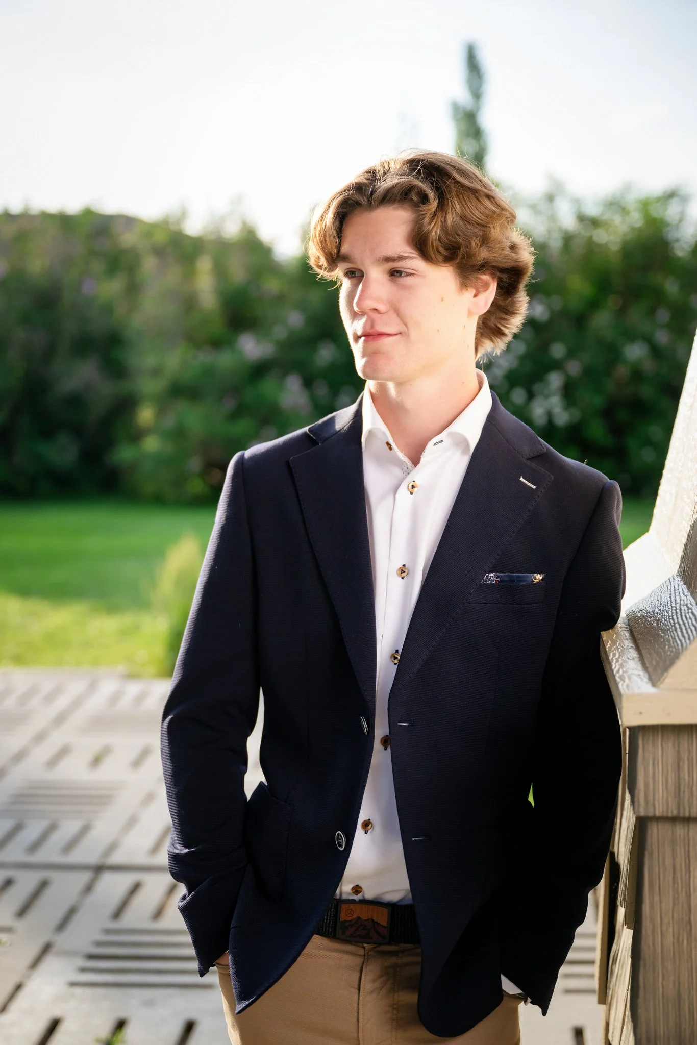 Young man with wavy brown hair leaning against a wooden railing outdoors, wearing a dark blazer and white shirt, with trees and a bright sky in the background.