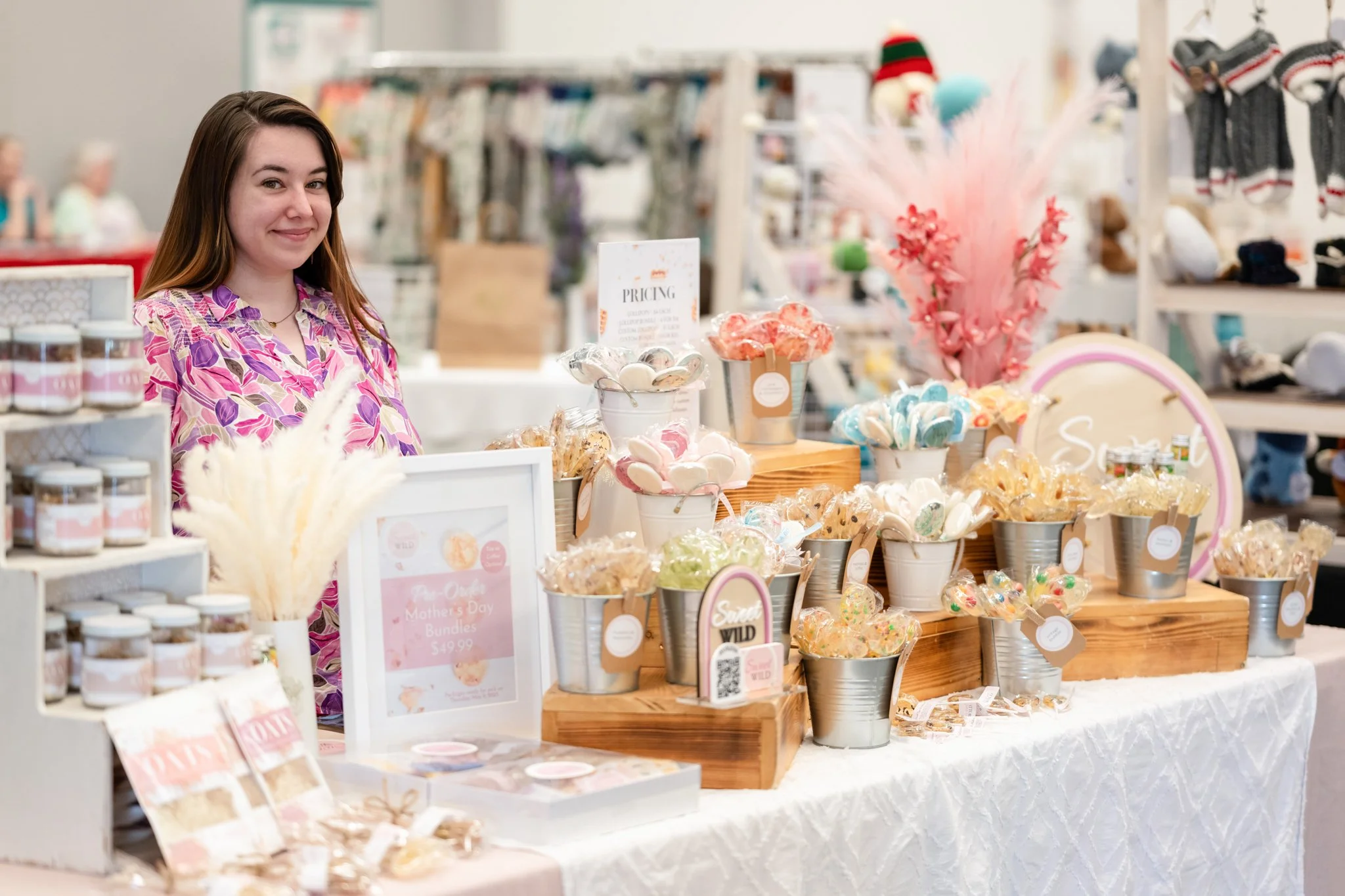 A woman standing behind a display table with Mother's Day themed items including jars, cookies, and decorative flowers at a market or craft fair.