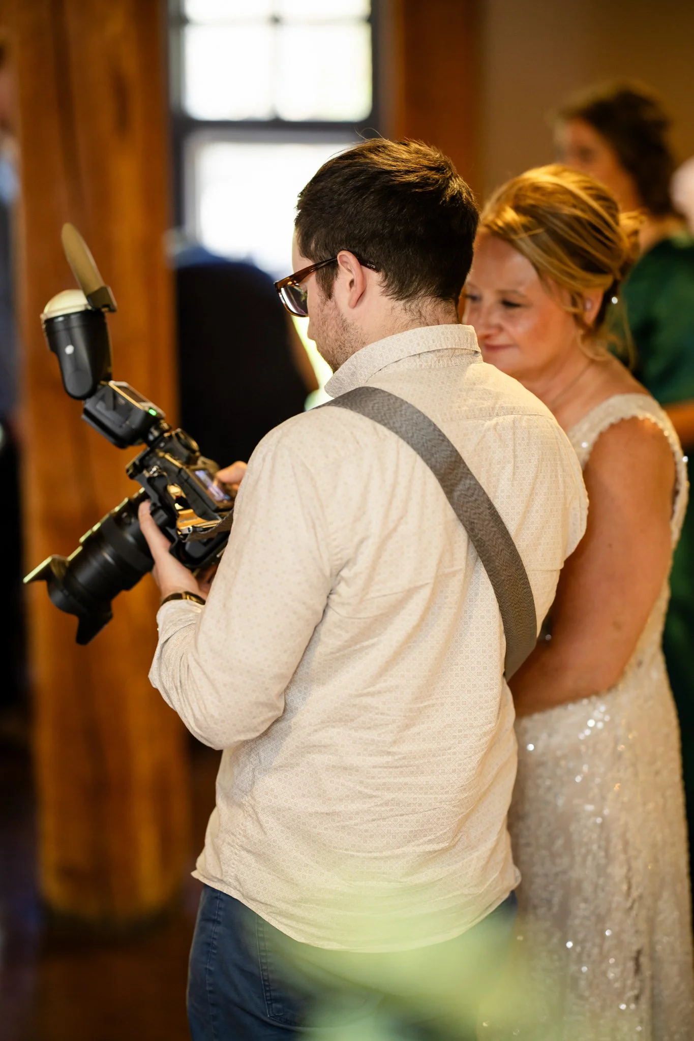 A photographer with glasses and a beige shirt standing while taking pictures at a wedding ceremony, with a woman in a white lace dress standing behind him.