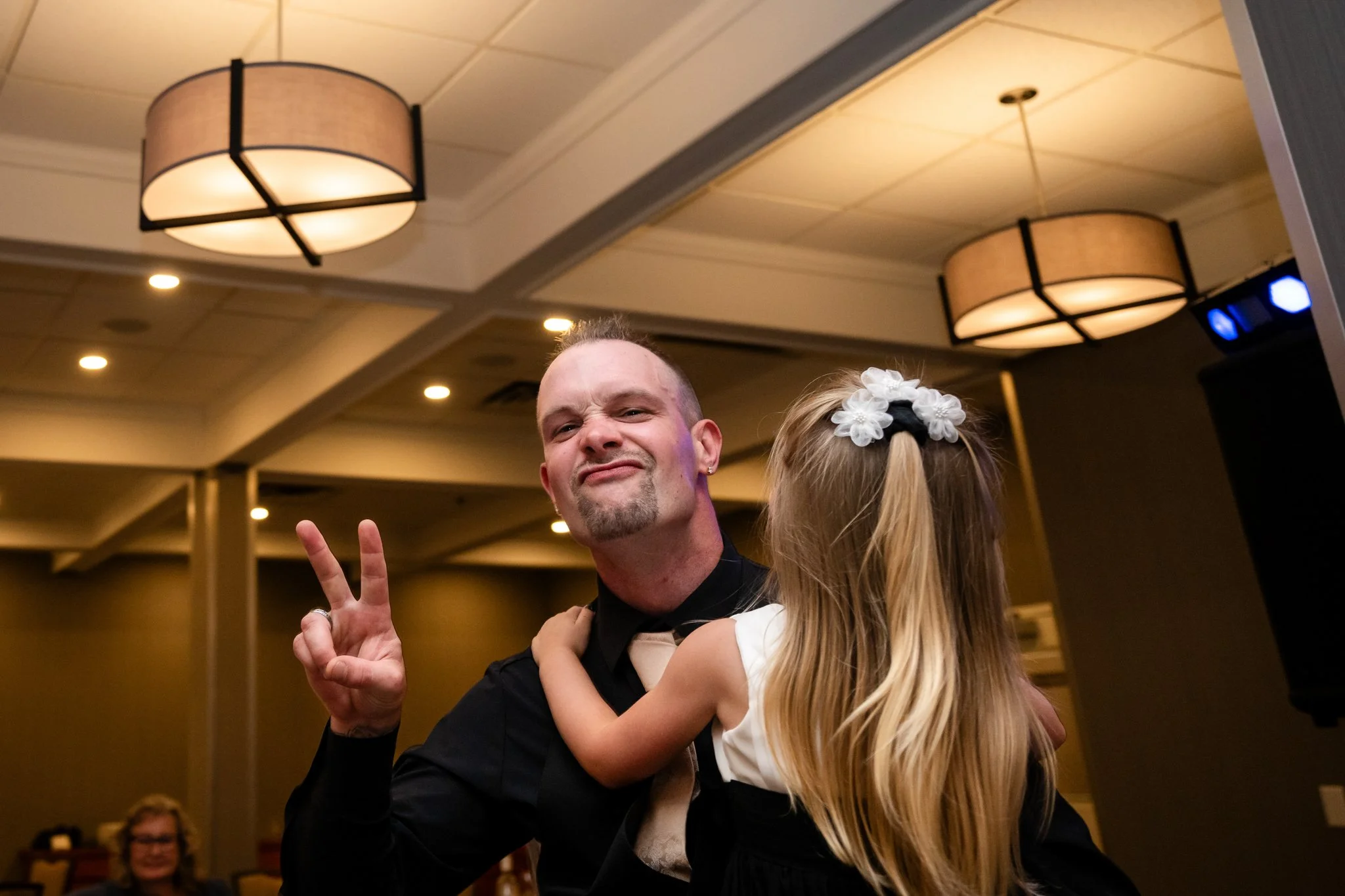 Man posing with peace sign while holding a young girl with long blonde hair in a white dress and floral hair accessory, at an indoor event.