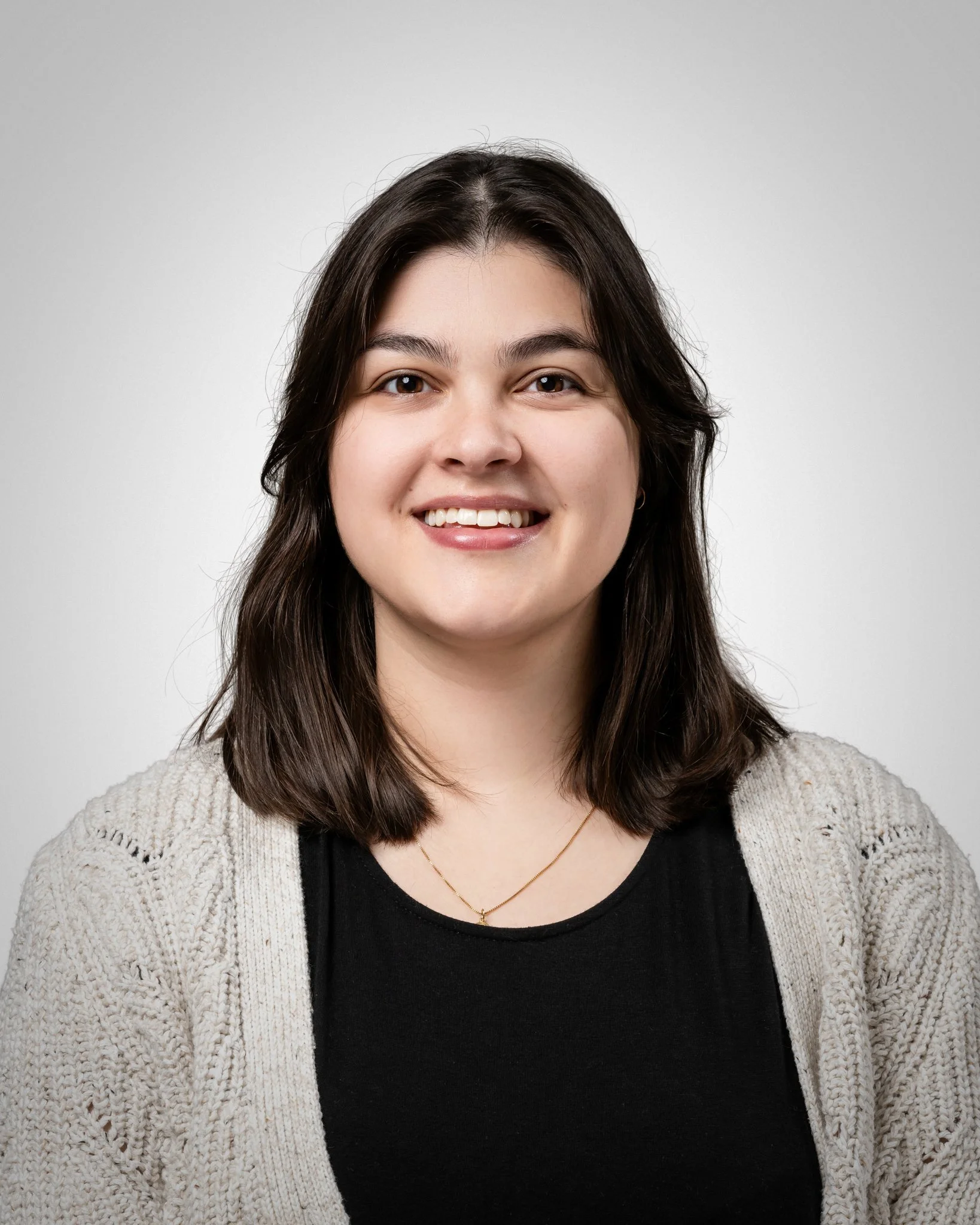 A young woman with shoulder-length brown hair smiling at the camera against a plain light gray background, wearing a black shirt, a cream-colored knitted cardigan, and a delicate gold necklace.