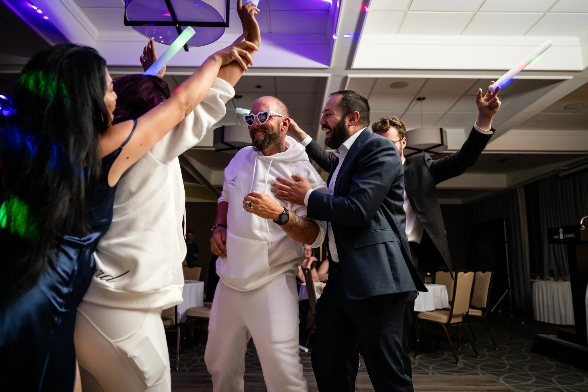 Group of people celebrating and dancing at a party or wedding reception, holding glow sticks, with one person wearing novelty sunglasses.