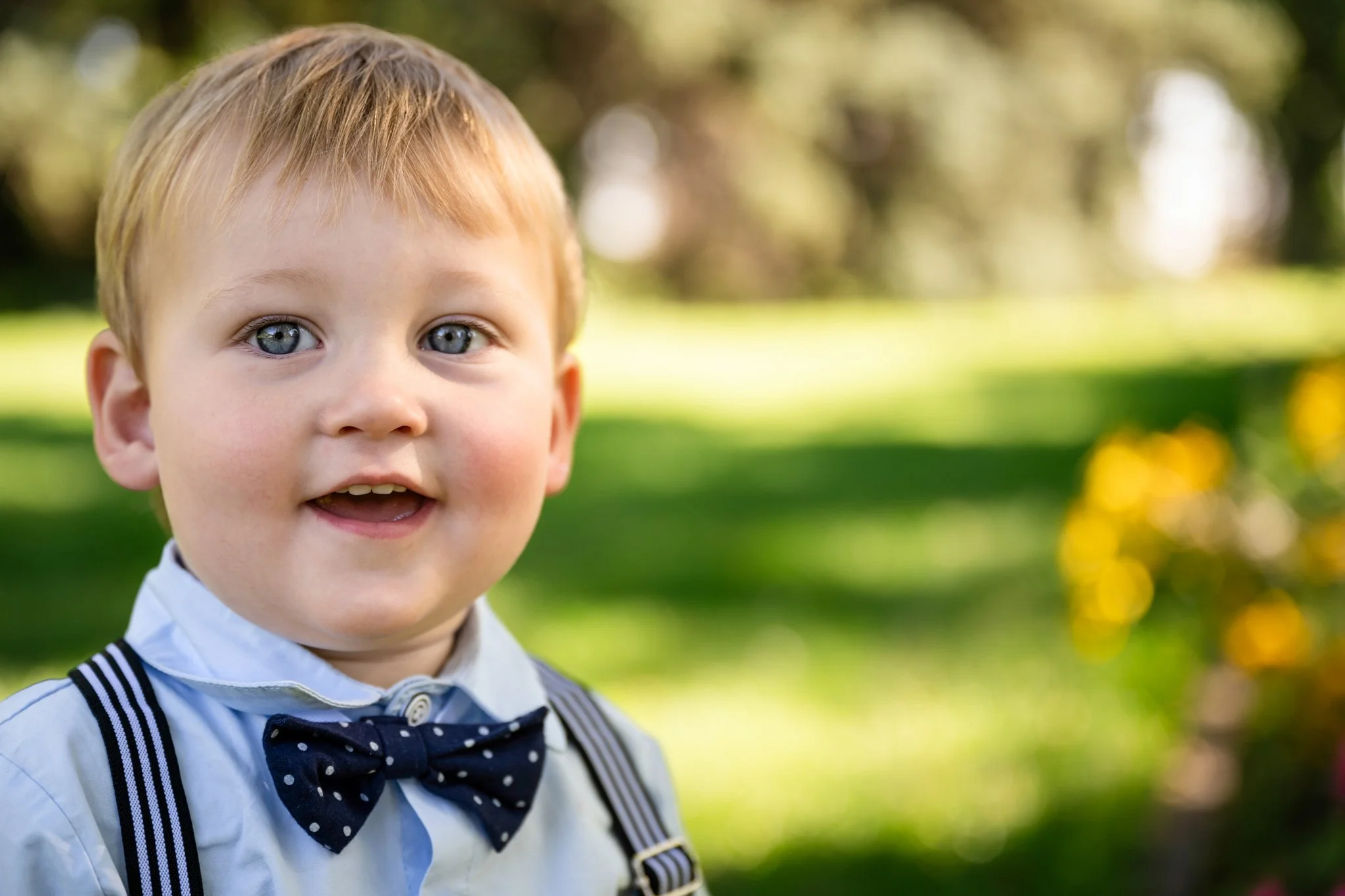 A smiling young boy with blonde hair wearing a white shirt, a polka-dotted bow tie, and suspenders, outdoors in a green park with trees and yellow flowers in the background.