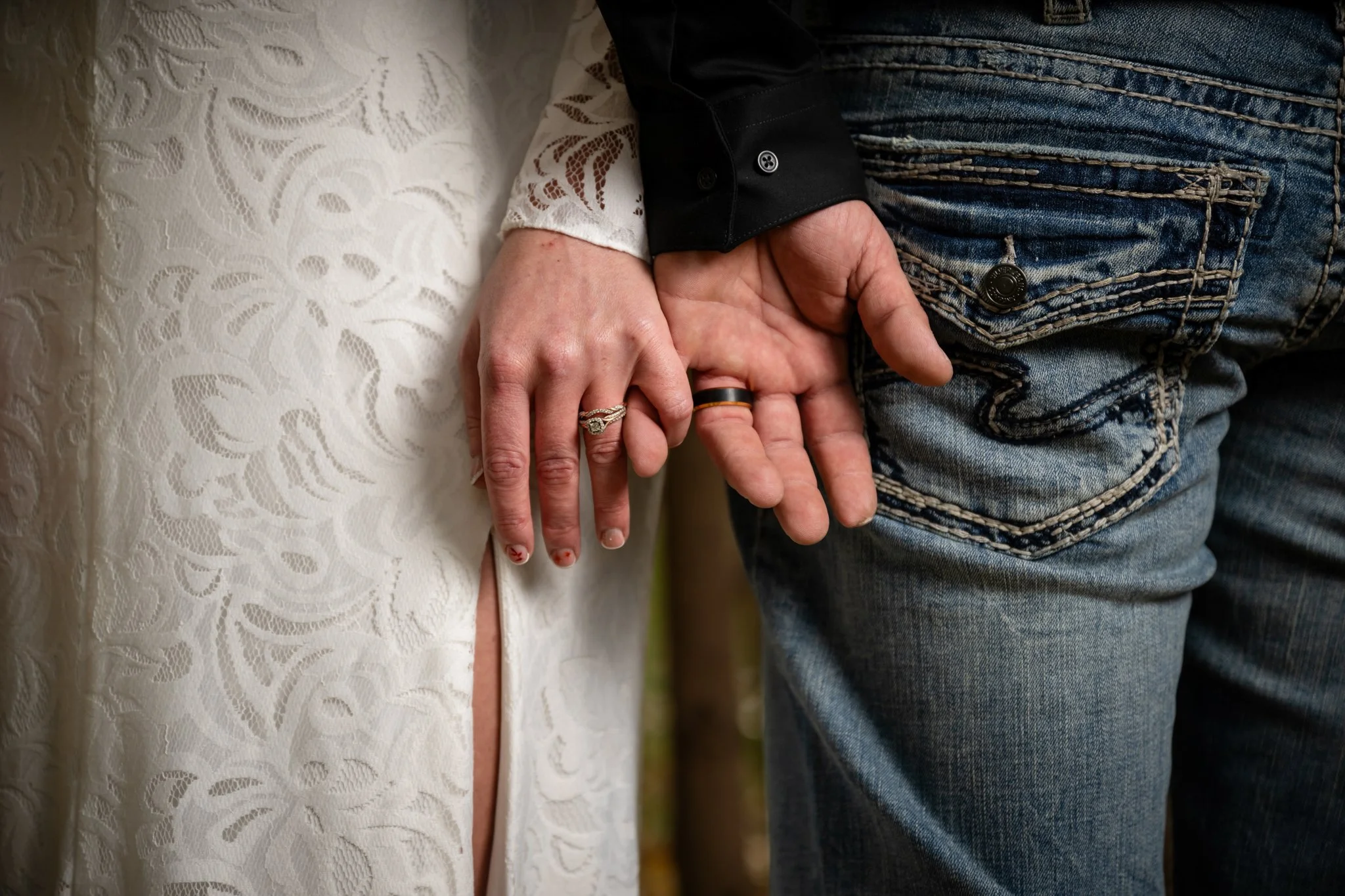 Close-up of a couple holding hands, showing wedding rings, with wedding dress and jeans.
