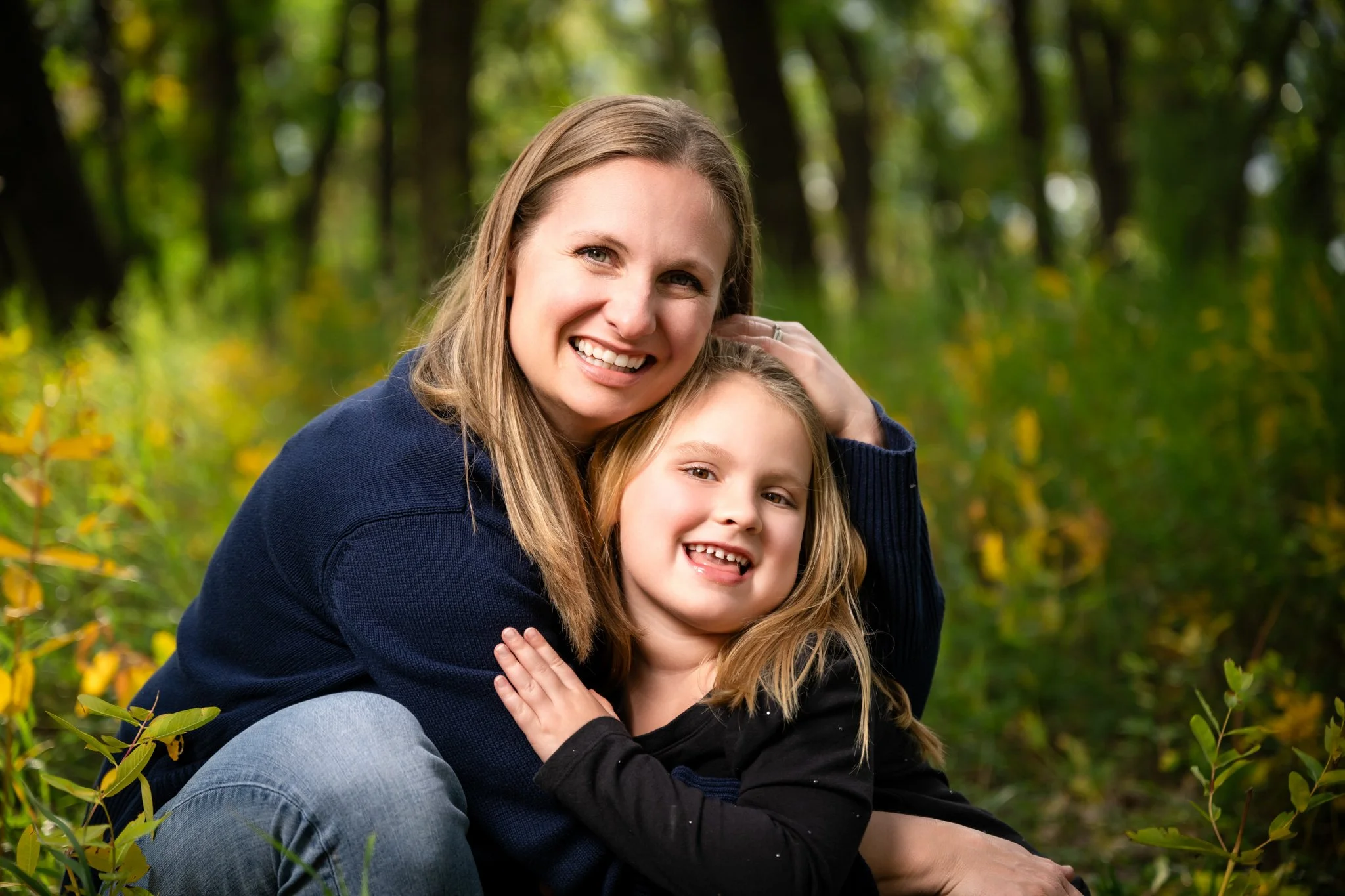 A woman and young girl smiling and hugging outdoors in a wooded area