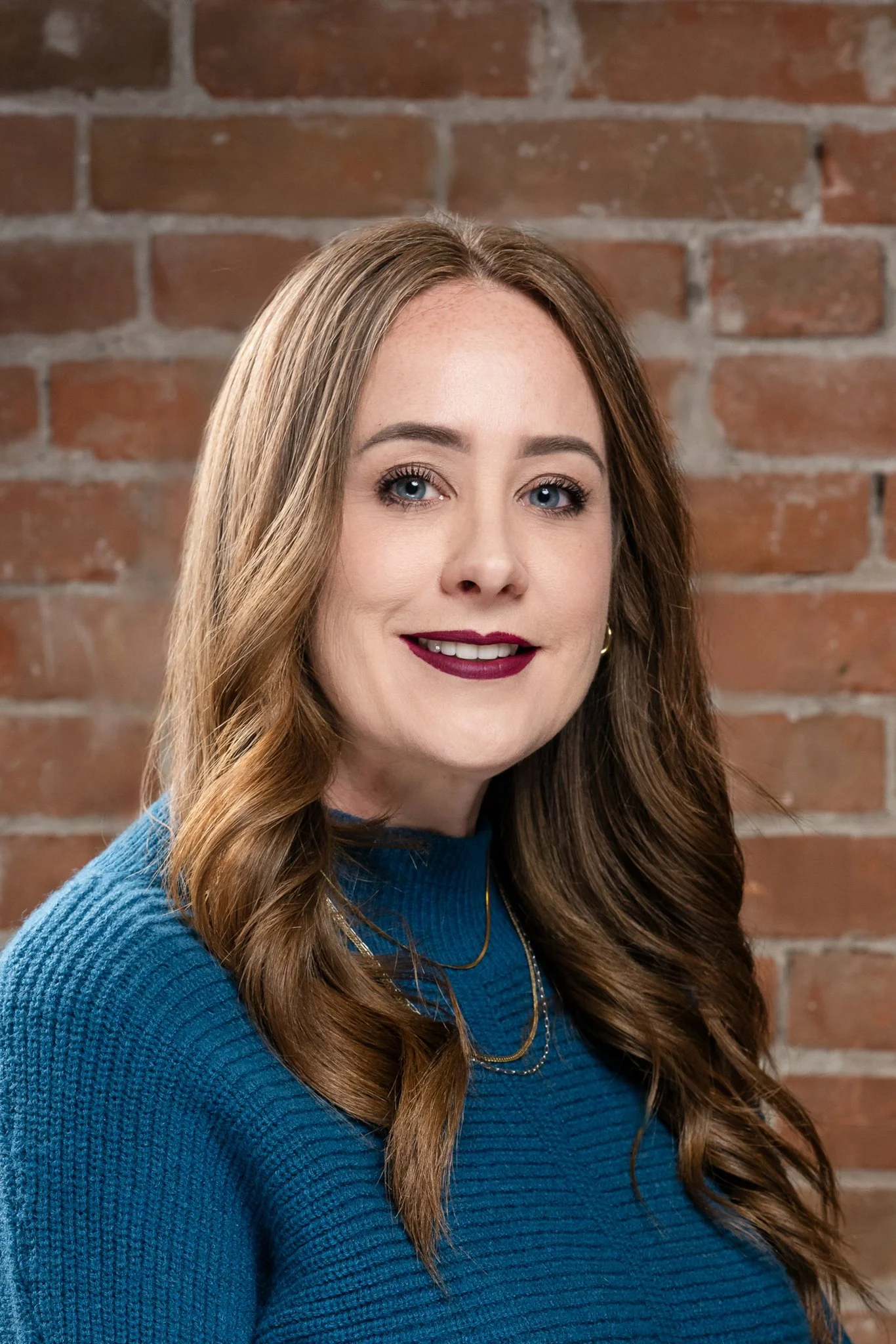 A woman with long wavy brown hair, blue eyes, wearing a teal sweater and gold jewelry, standing against a brick wall.