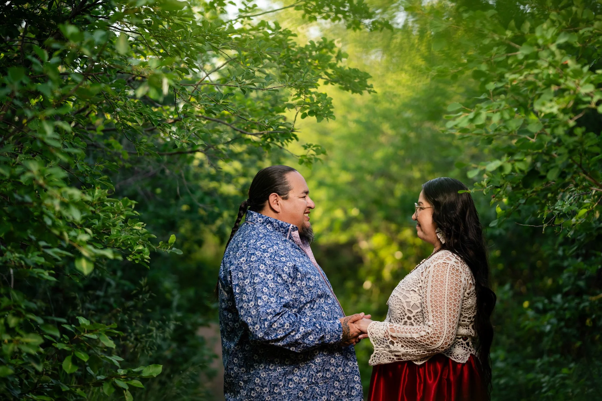 A couple standing outdoors in a green, leafy setting, holding hands and smiling at each other.