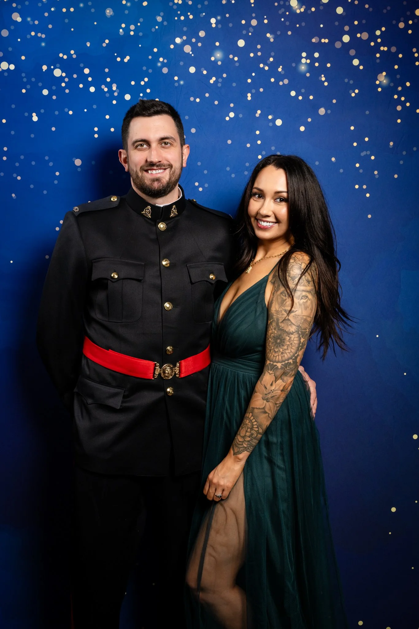 A man in a formal black military uniform with a red belt standing next to a woman in a dark green gown with a thigh-high slit, both smiling against a blue background with gold confetti.