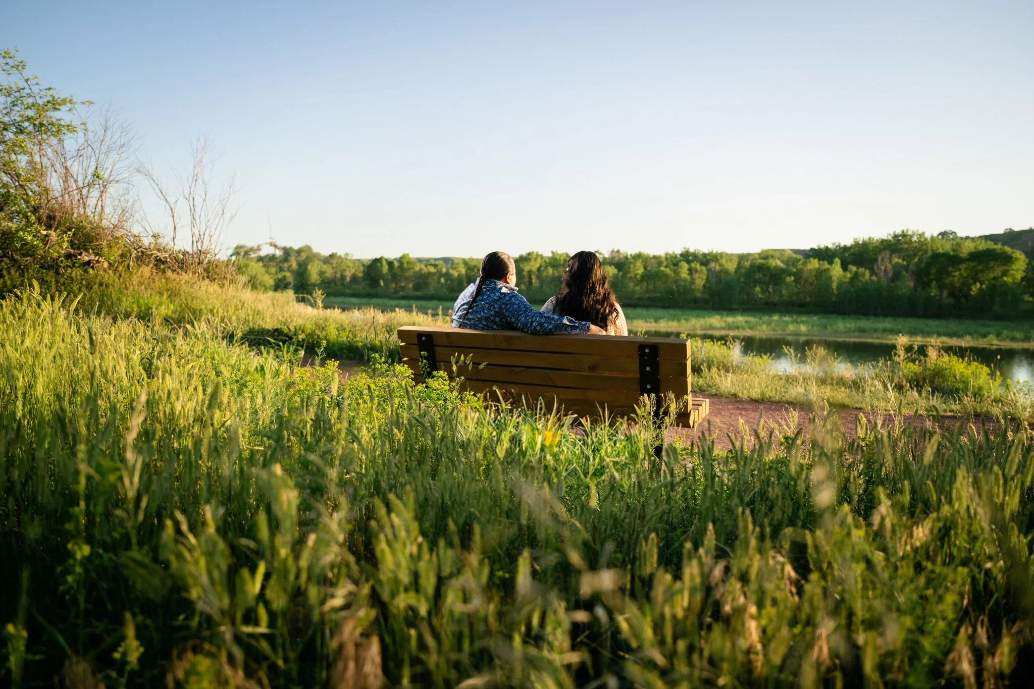 Two women sitting on a park bench overlooking a river, surrounded by green grass and trees, on a sunny day.