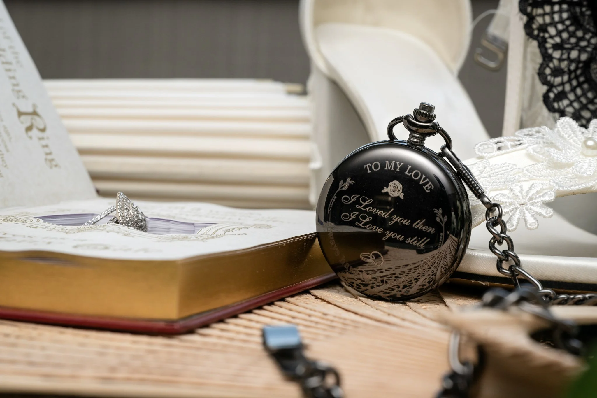 A black pocket watch with an inscription reading 'To My Love, I loved you then, I love you still' resting on an open book with a diamond engagement ring on its pages.